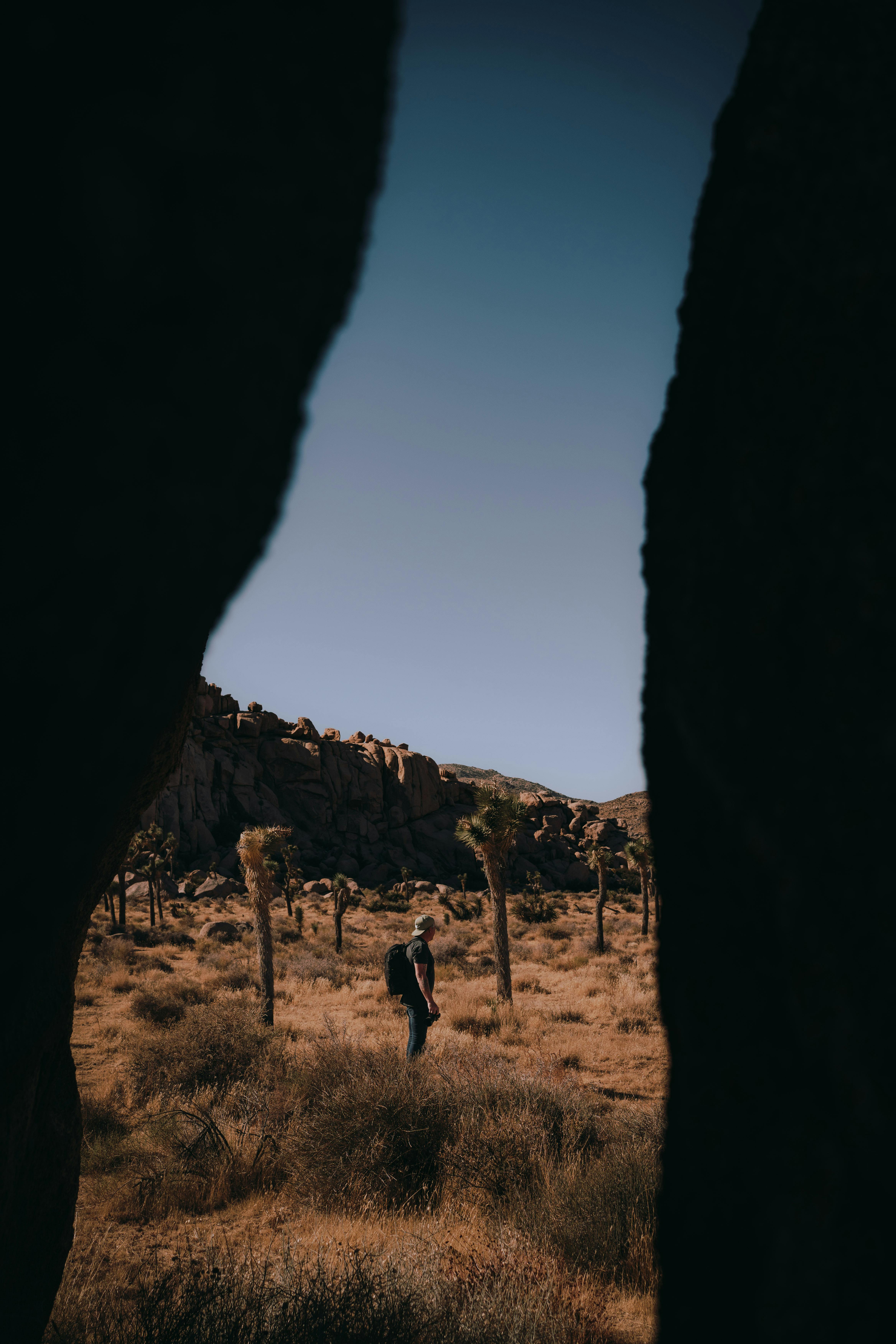 A lone hiker ventures through a desert landscape framed by large rocks, with dry shrubs and clear skies.