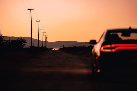 Silhouette of a car driving on a dirt road with sunset hues and distant mountains.