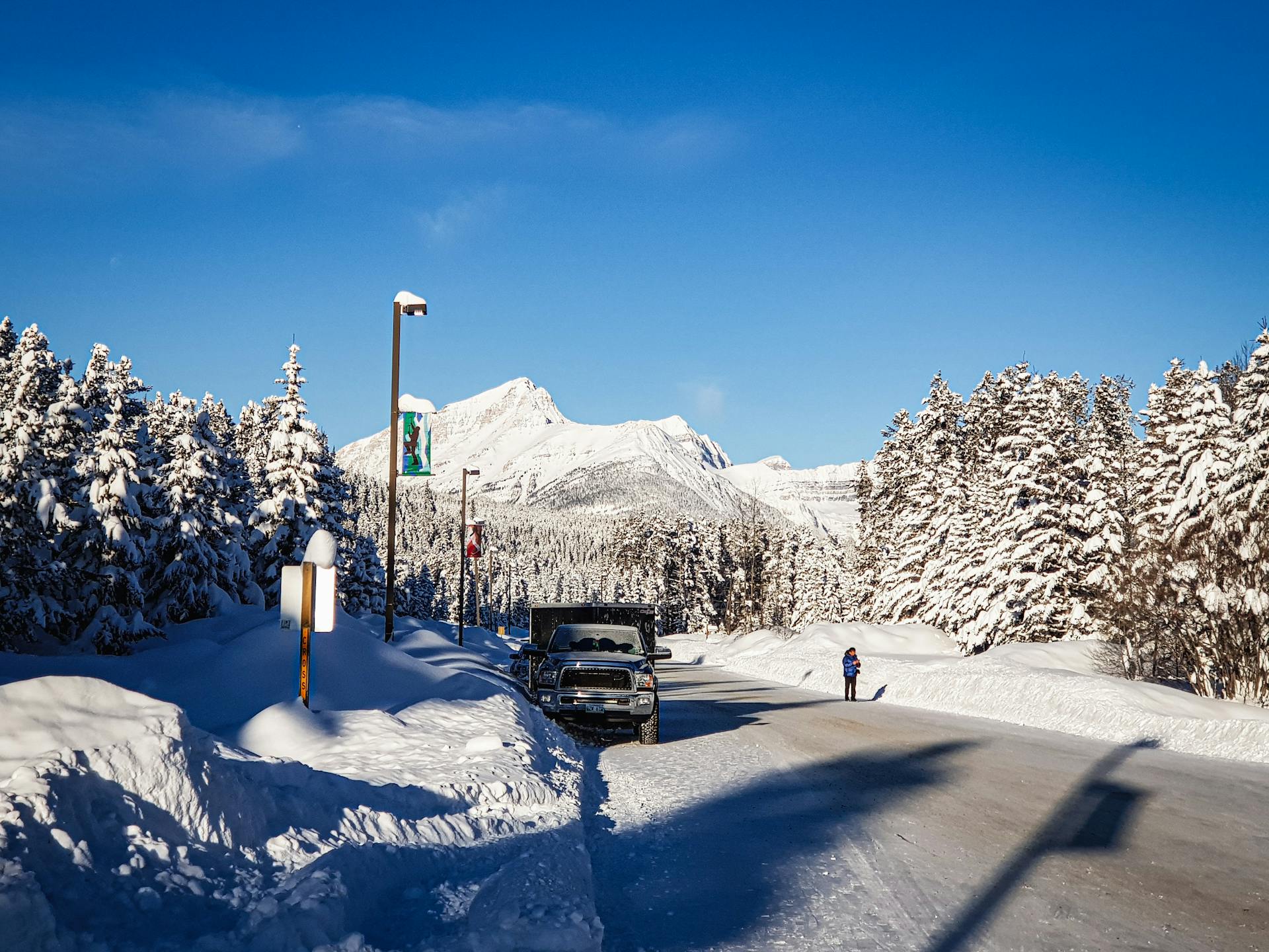 A winter scene in Canada featuring a snow-covered road, truck, and person in a mountainous forest area.