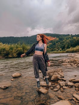 A woman enjoys a peaceful hike along a scenic river with lush forests and mountains.