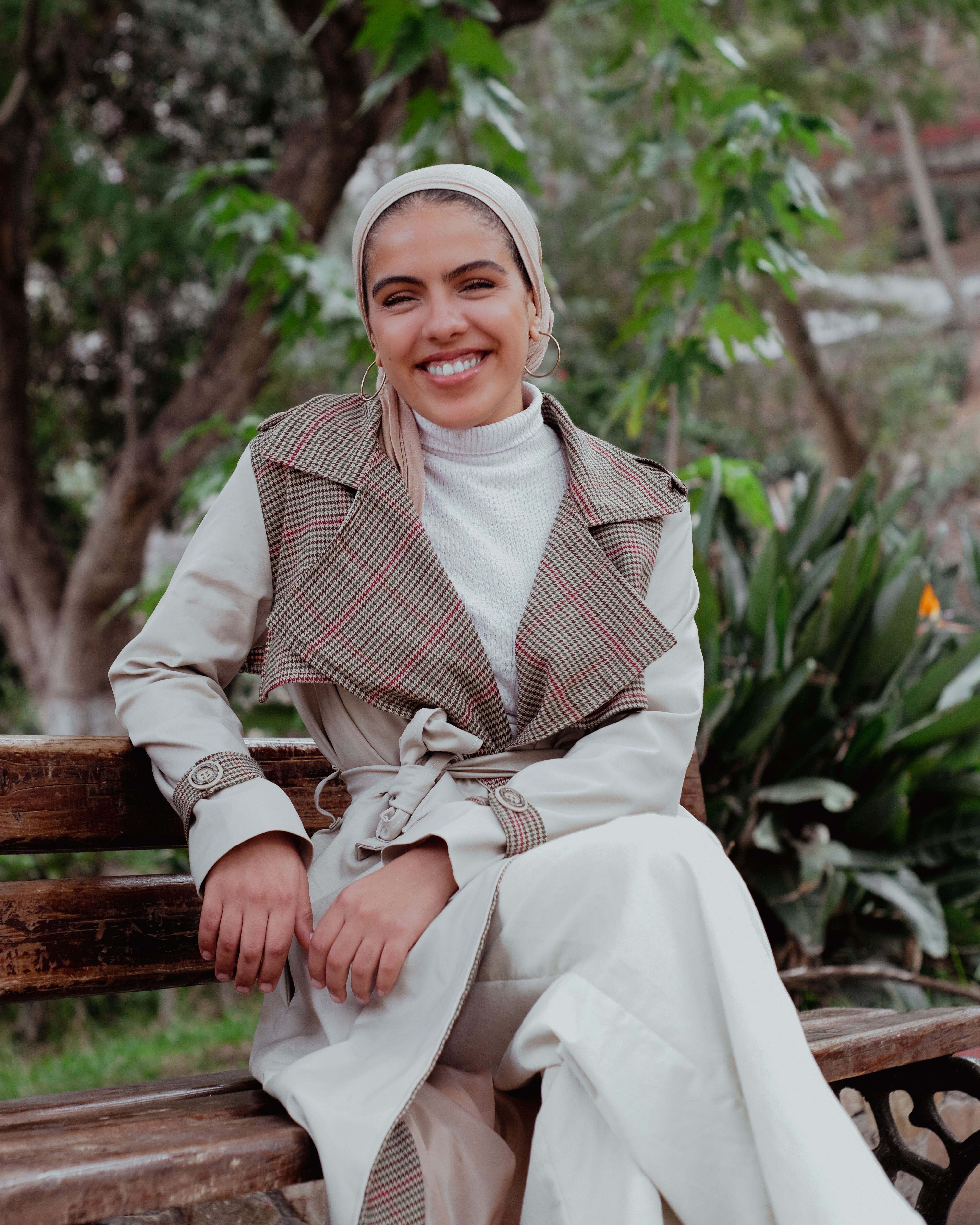 A woman in a fashionable trench coat smiles while sitting on a park bench outdoors.