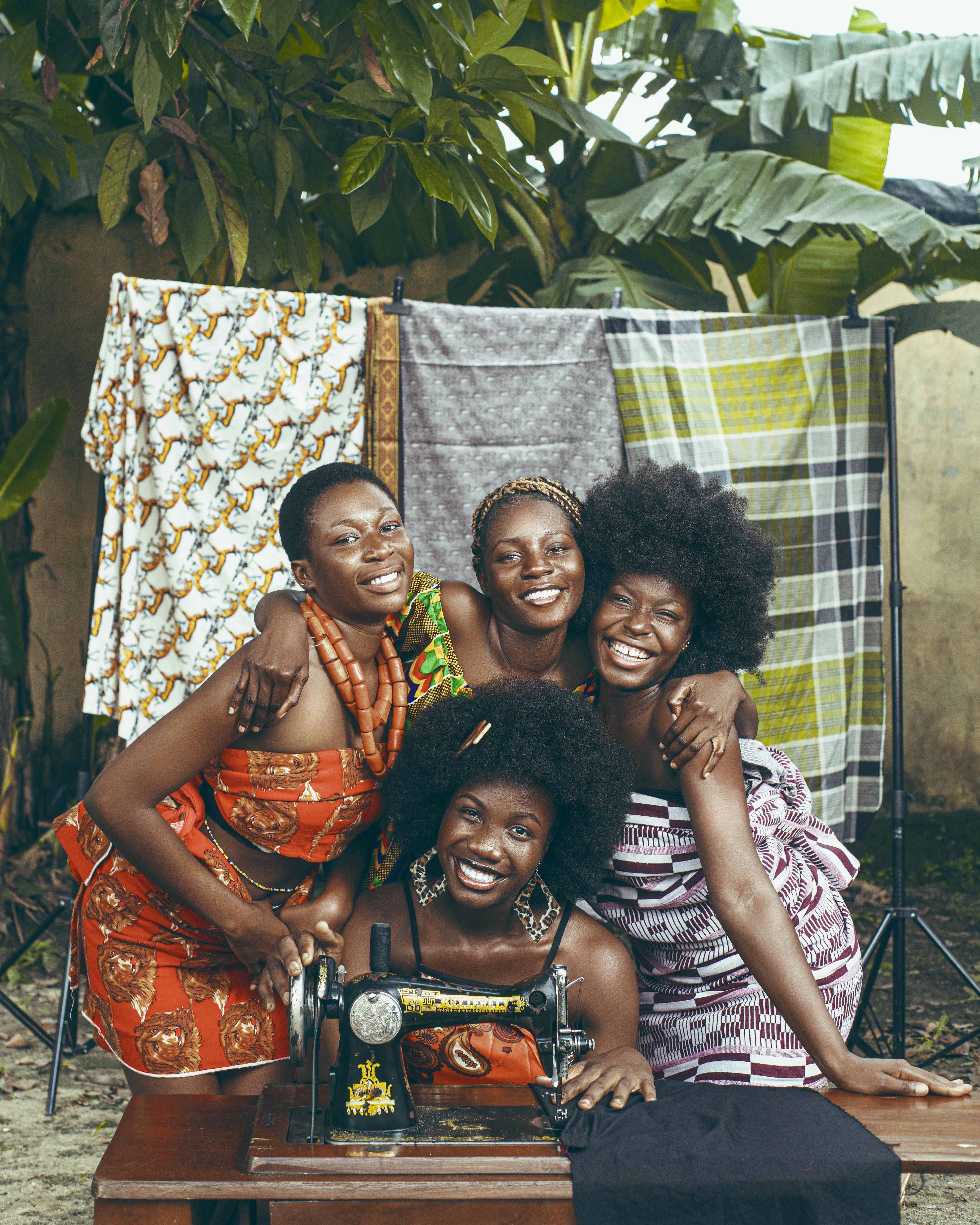 Group of happy African women in colorful fabrics posing with a sewing machine outside.