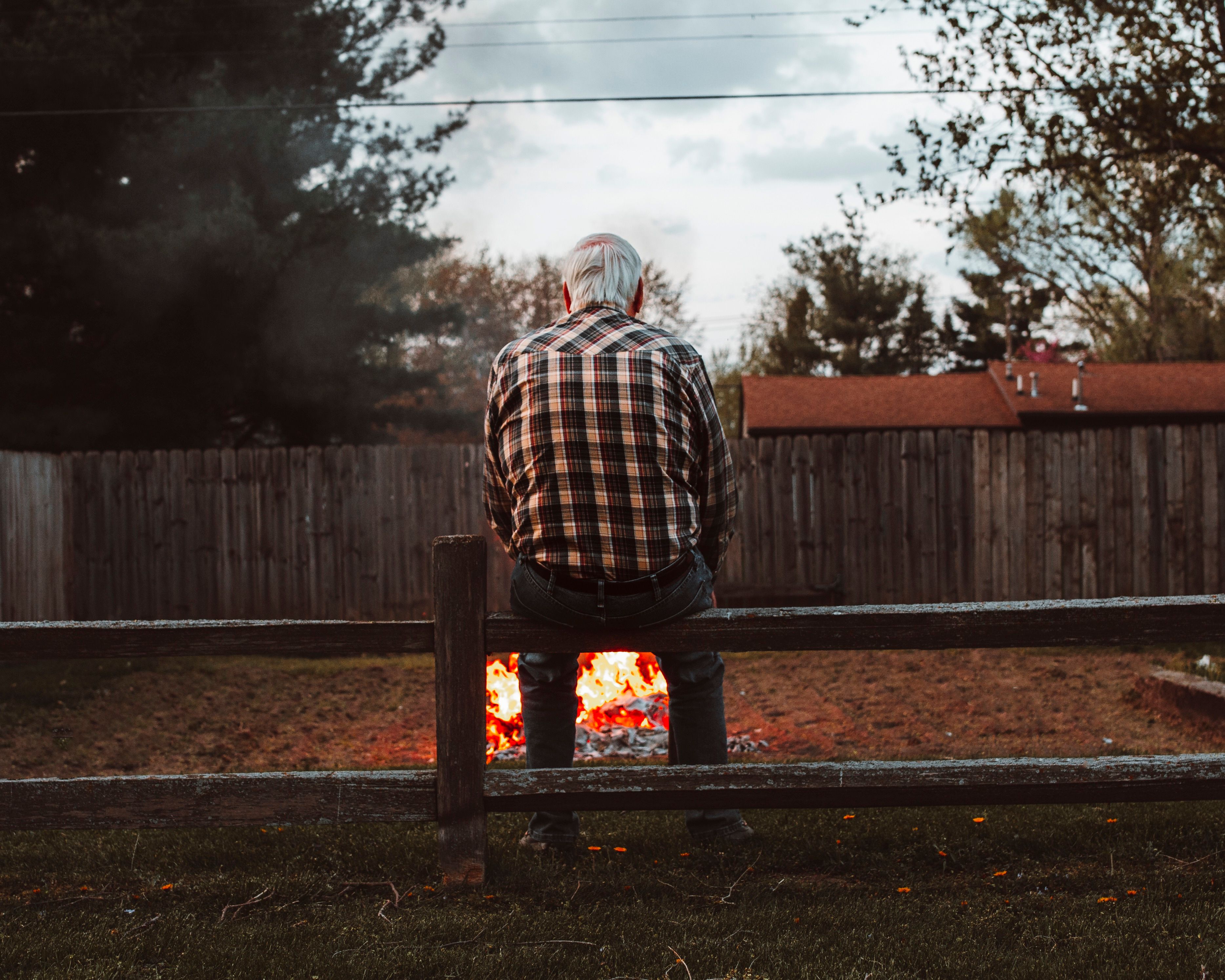 A senior man enjoys a peaceful evening sitting by a bonfire in a fenced backyard.