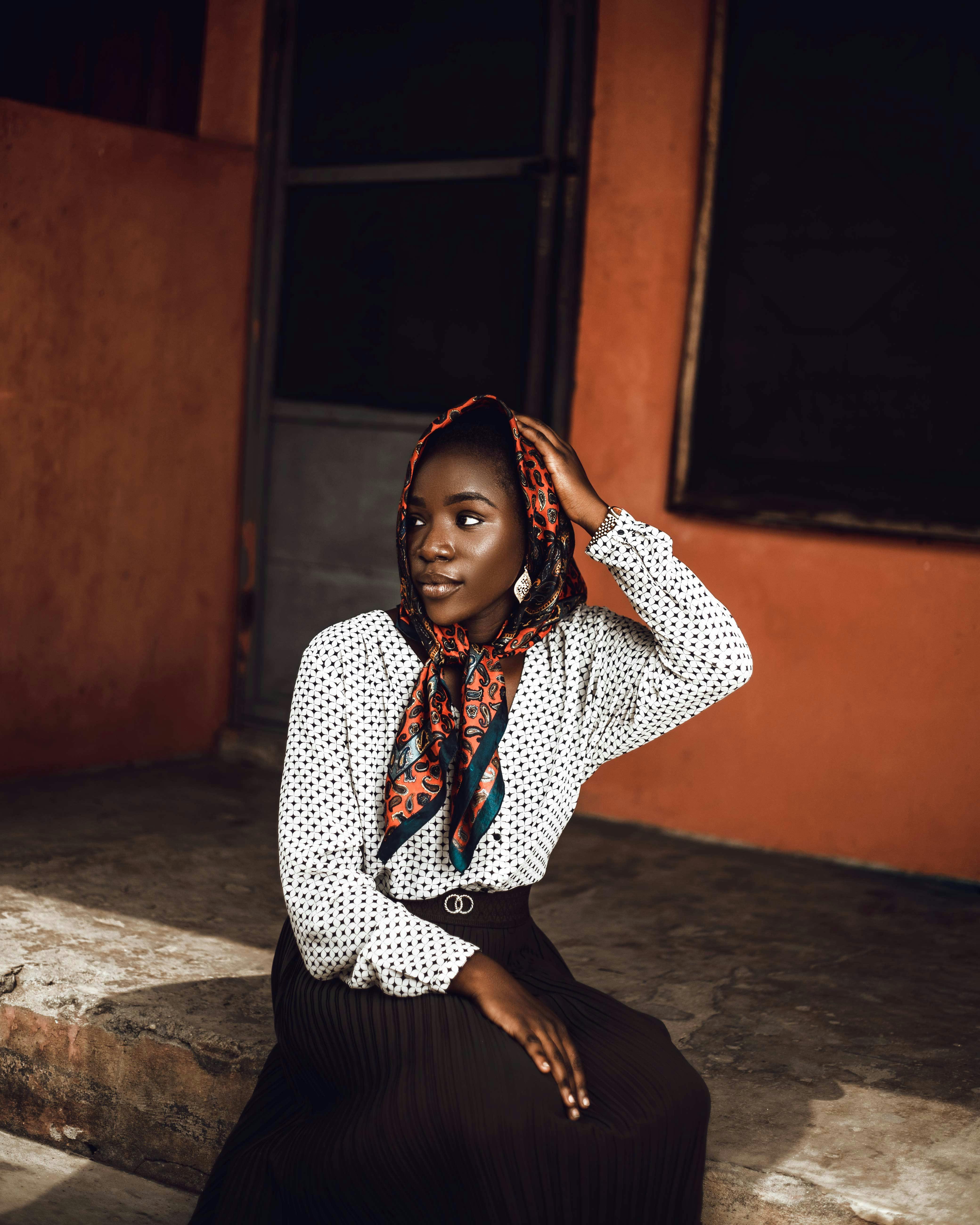 Stylish young African woman in polka dot blouse and headscarf sitting elegantly outdoors.