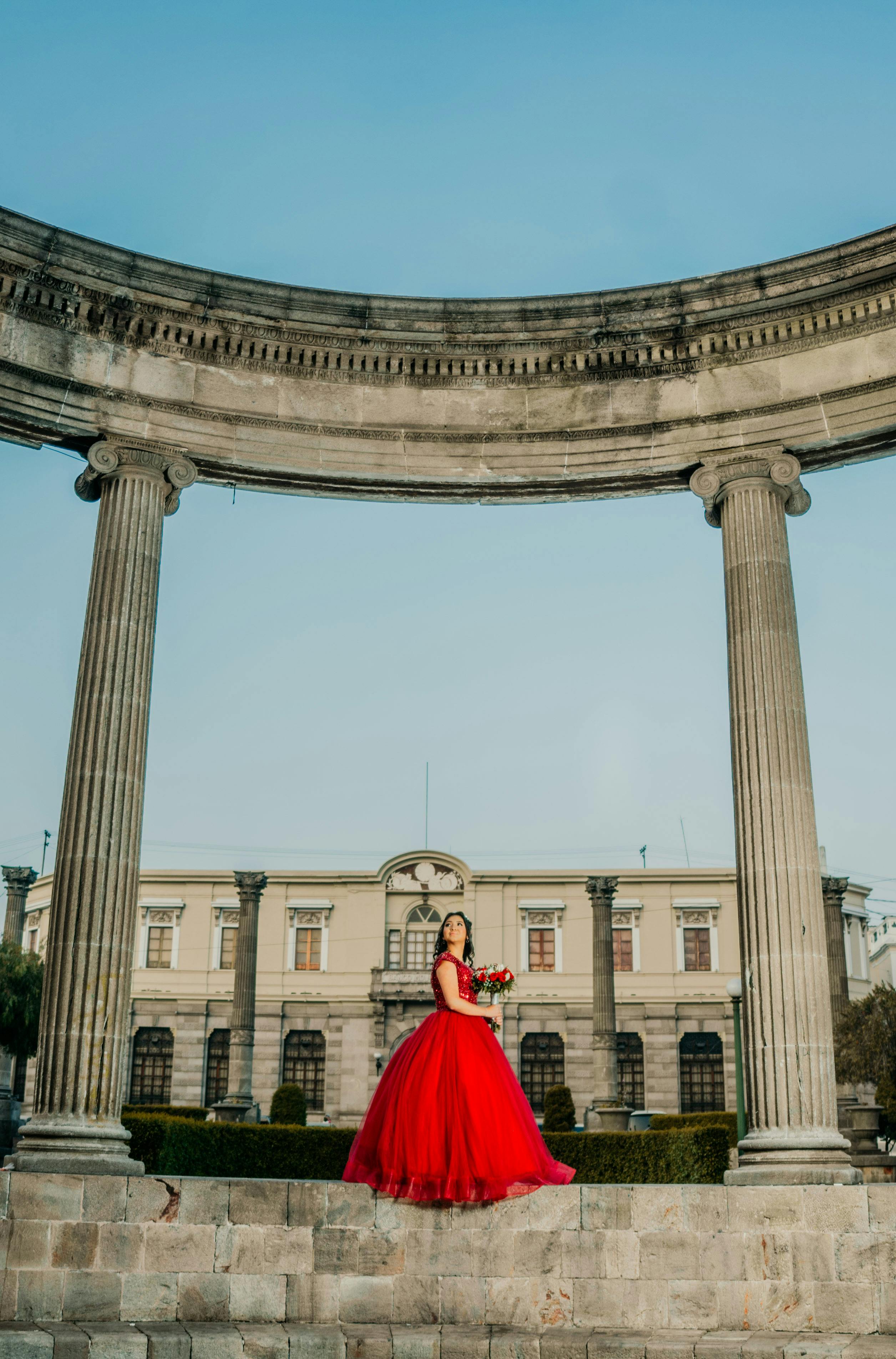 Woman in a red dress stands elegantly between ancient columns of a historic monument.