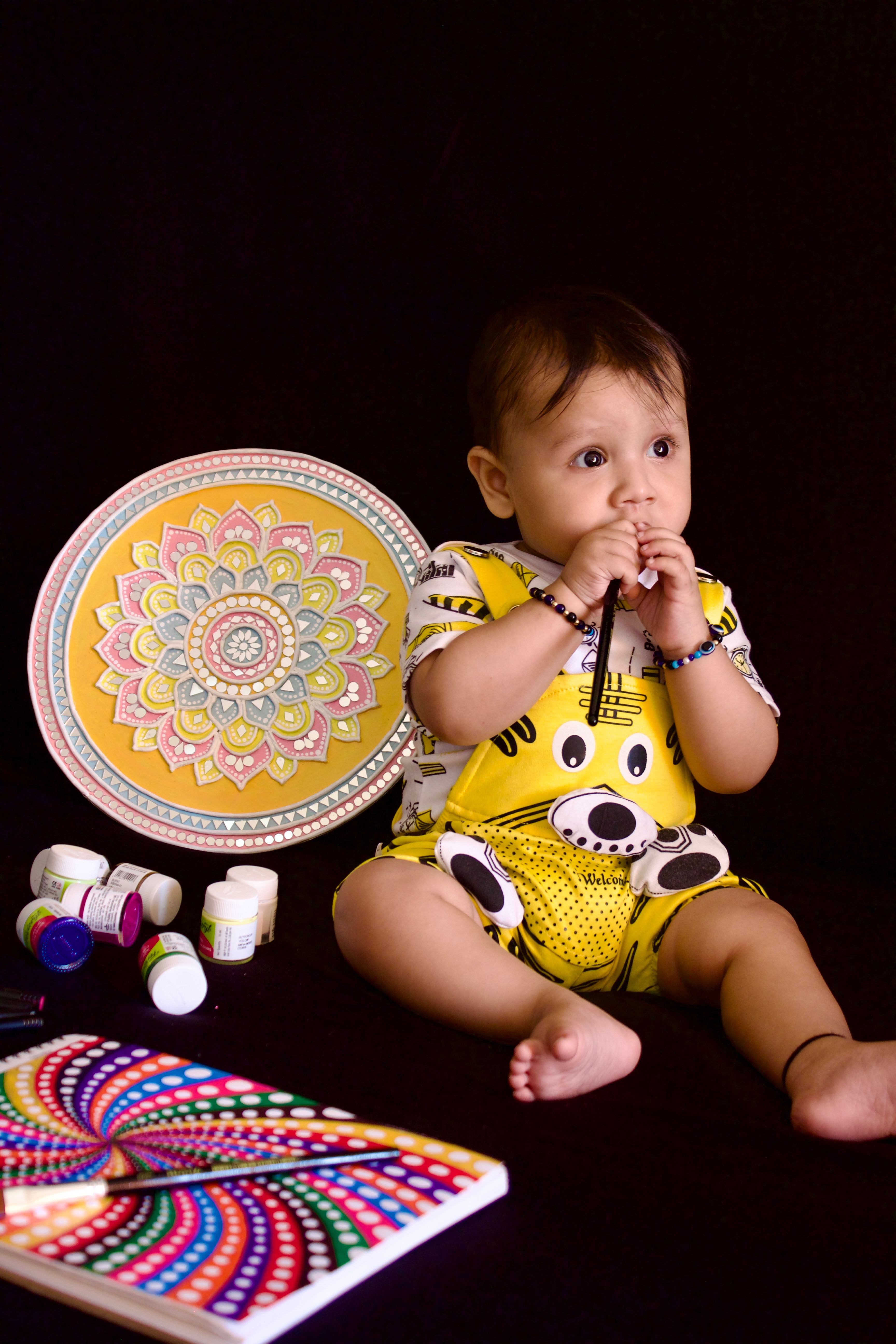 Cute baby in colorful overalls with art supplies and mandala art on black background.