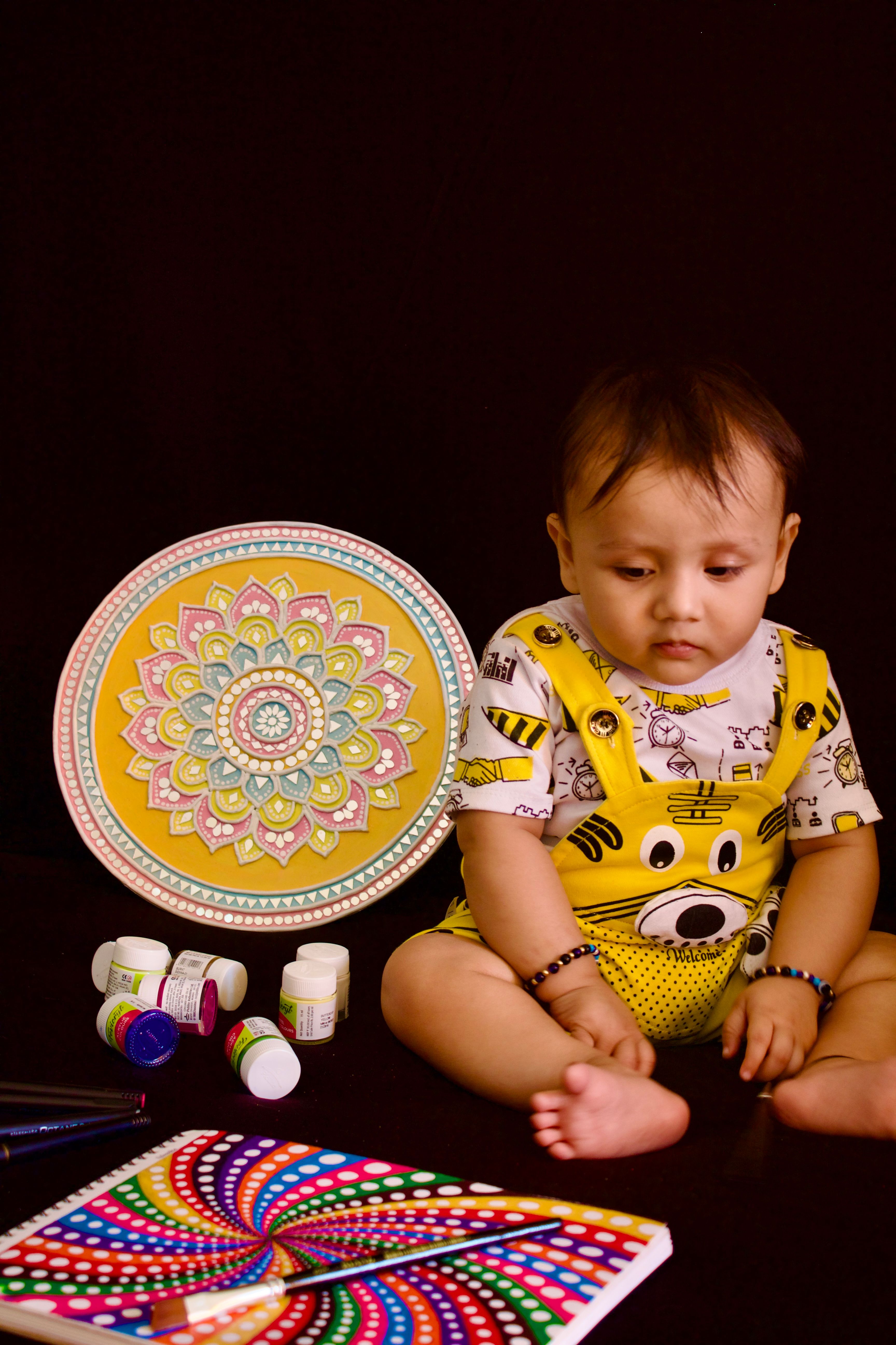 Adorable baby in yellow dungarees sitting with colorful mandala art and paints.