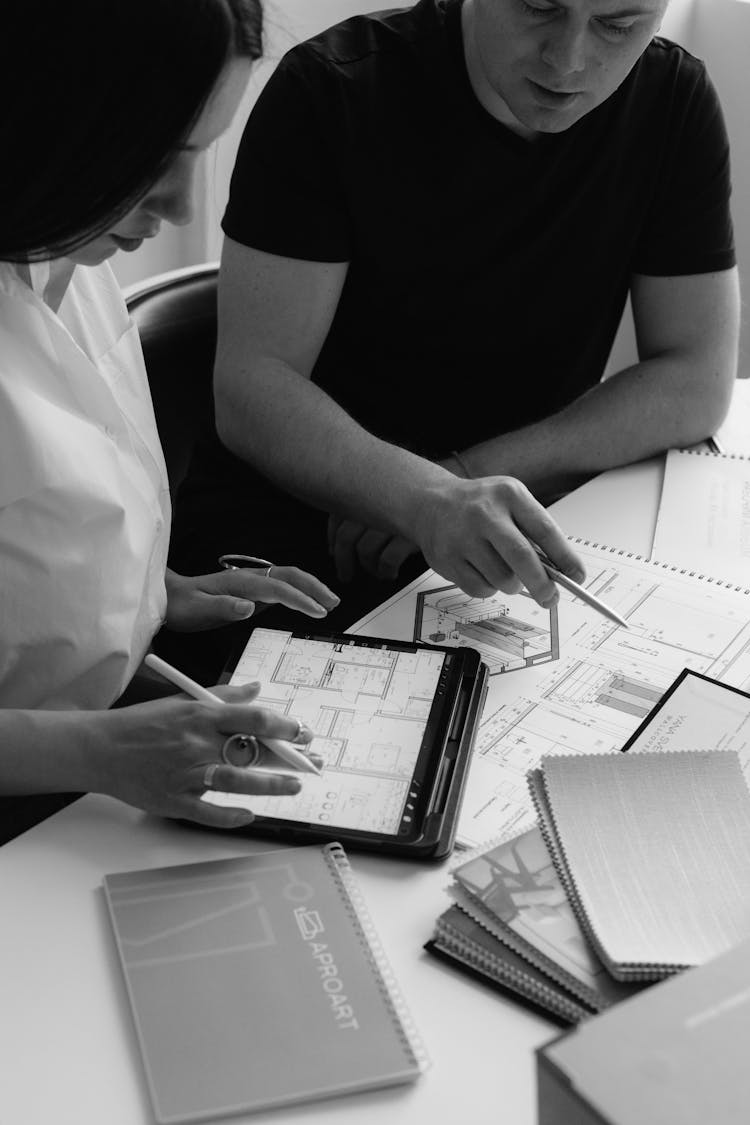 Man And Woman Sitting At The Desk With Papers And A Tablet, Looking At Architectural Designs 