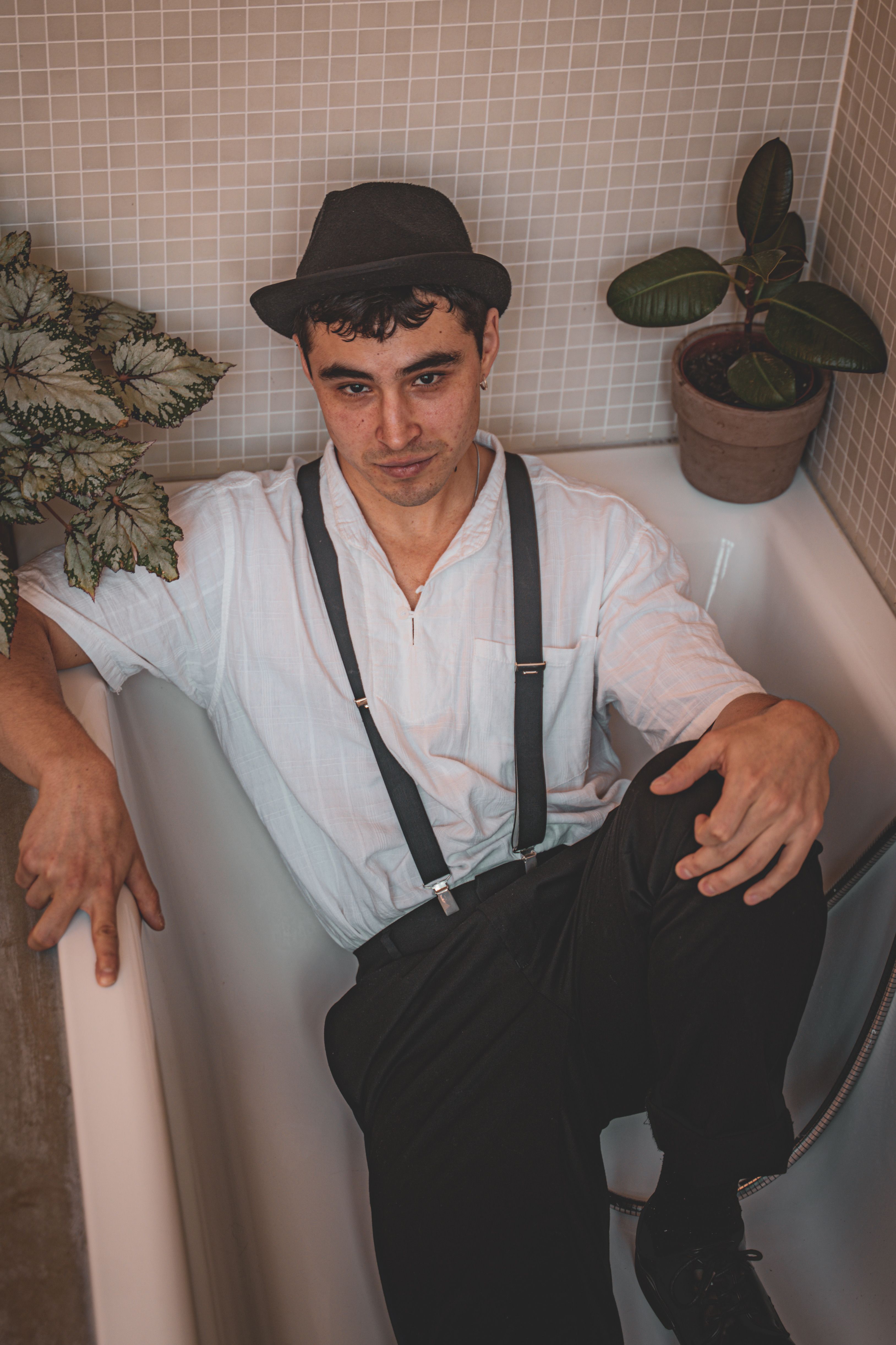 Stylish young man in a bathtub, wearing a hat and white shirt, surrounded by houseplants.