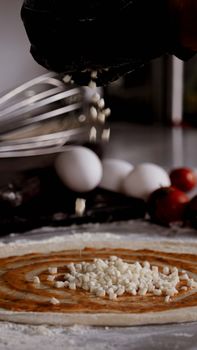 Close-up of pizza preparation, adding cheese with fresh ingredients on kitchen counter.