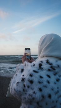 Woman in headscarf taking ocean photo with mobile at dawn.