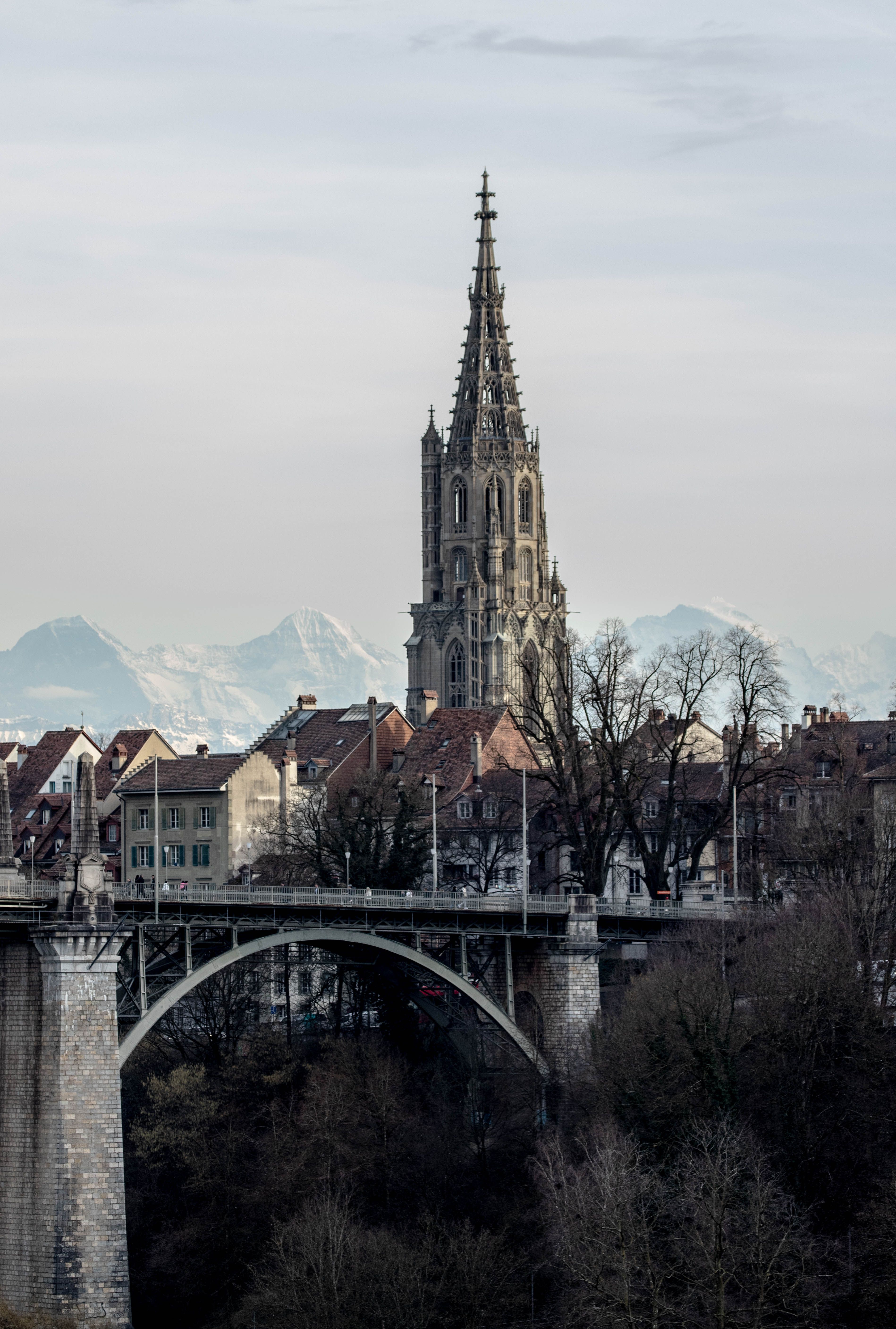 View of the Bern Cathedral and bridge with mountain backdrop, showcasing urban beauty.