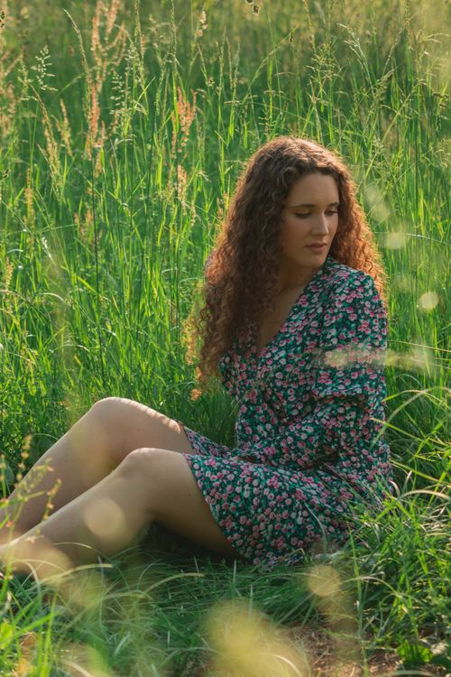 Free A woman with curly hair sits in a sunlit meadow in a floral dress, embodying tranquility and nature. Stock Photo