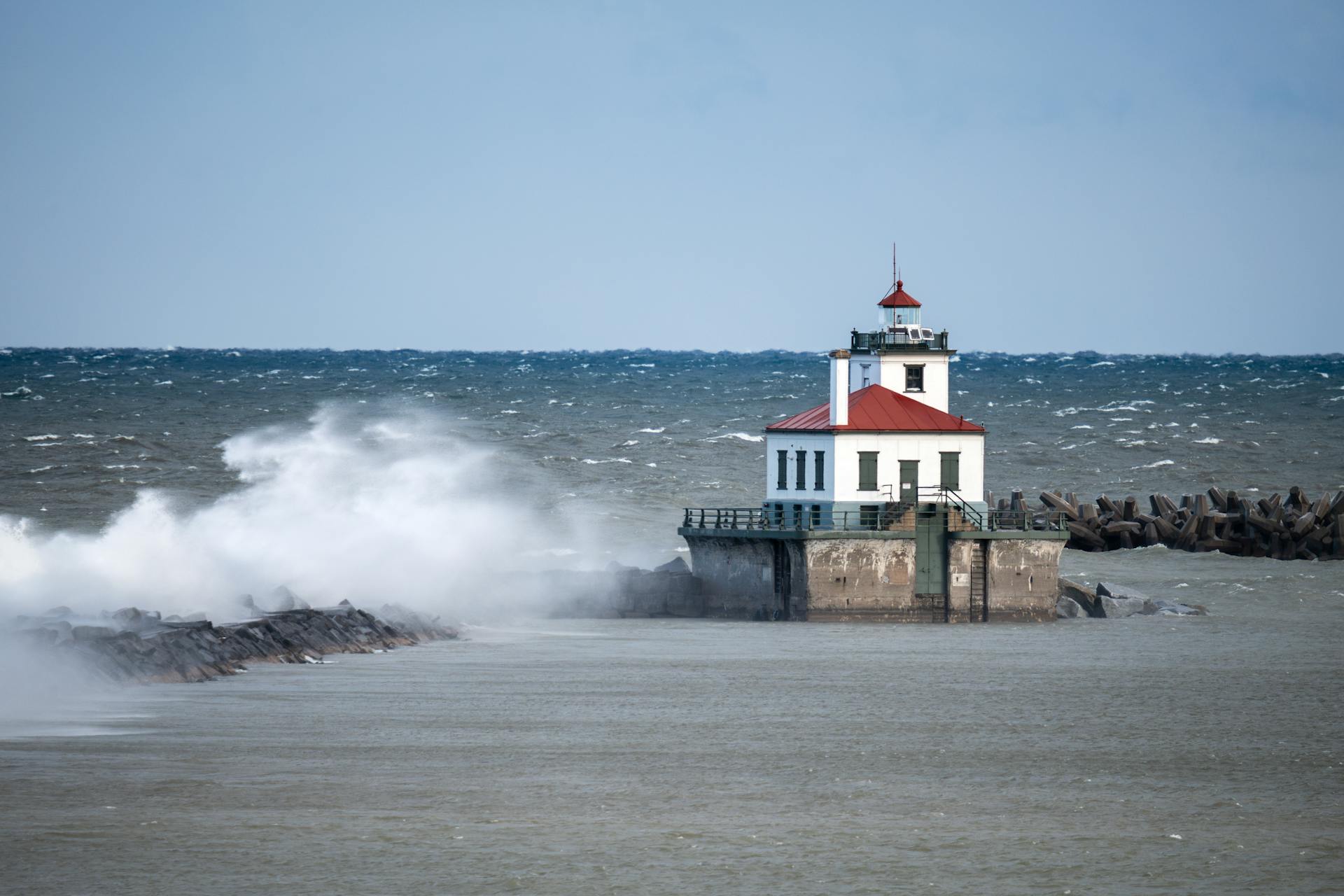Dramatic waves crash against the West Pierhead Lighthouse in Oswego, NY, under a clear blue sky.