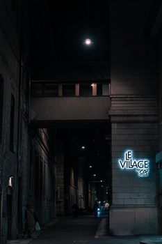 Urban alleyway at night featuring a neon 'Le Village' sign, creating a mysterious ambience.