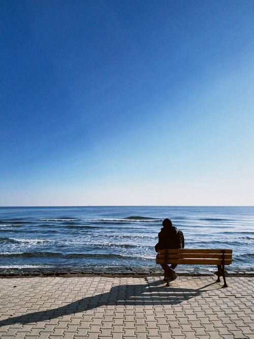 Free A person sits alone on a bench overlooking a calm sea under a clear sky, capturing a moment of solitude. Stock Photo
