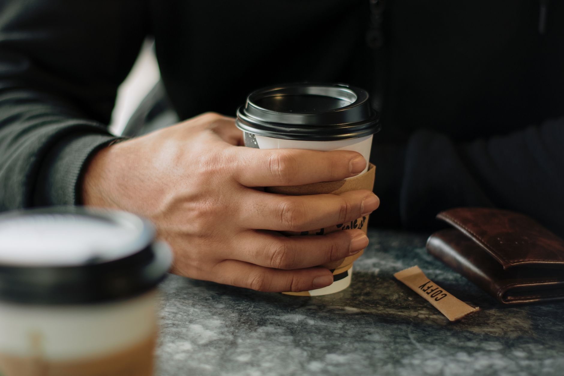 A close-up of a man's hand holding a to-go coffee cup inside a cafe.. close, mans, hand