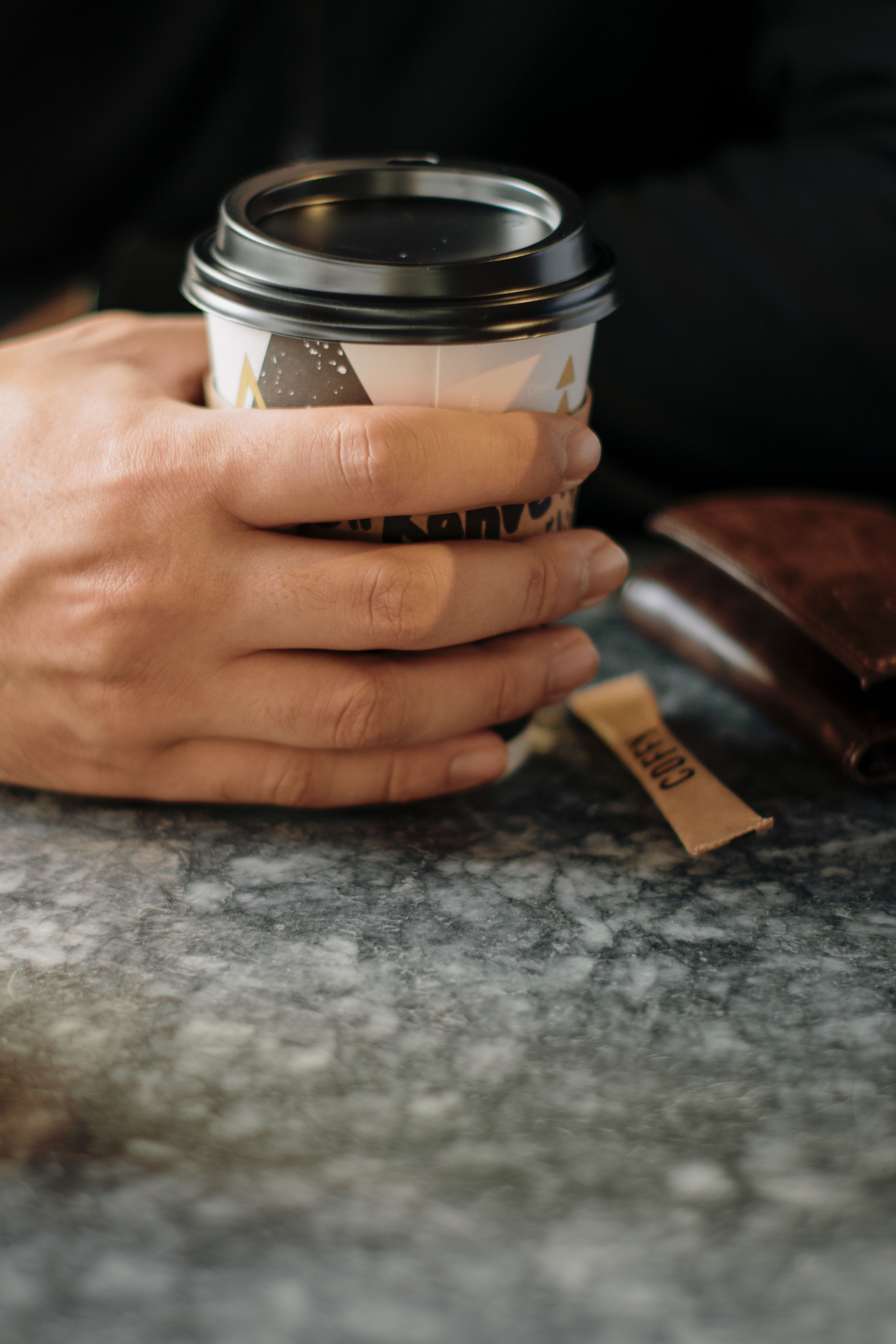 Close-up of a hand holding a coffee cup on a marble table with a brown wallet.. close, hand, holding