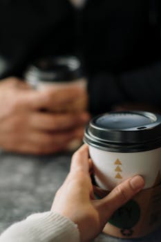 Two hands holding takeaway coffee cups in a cozy cafe setting, perfect for lifestyle and beverages.