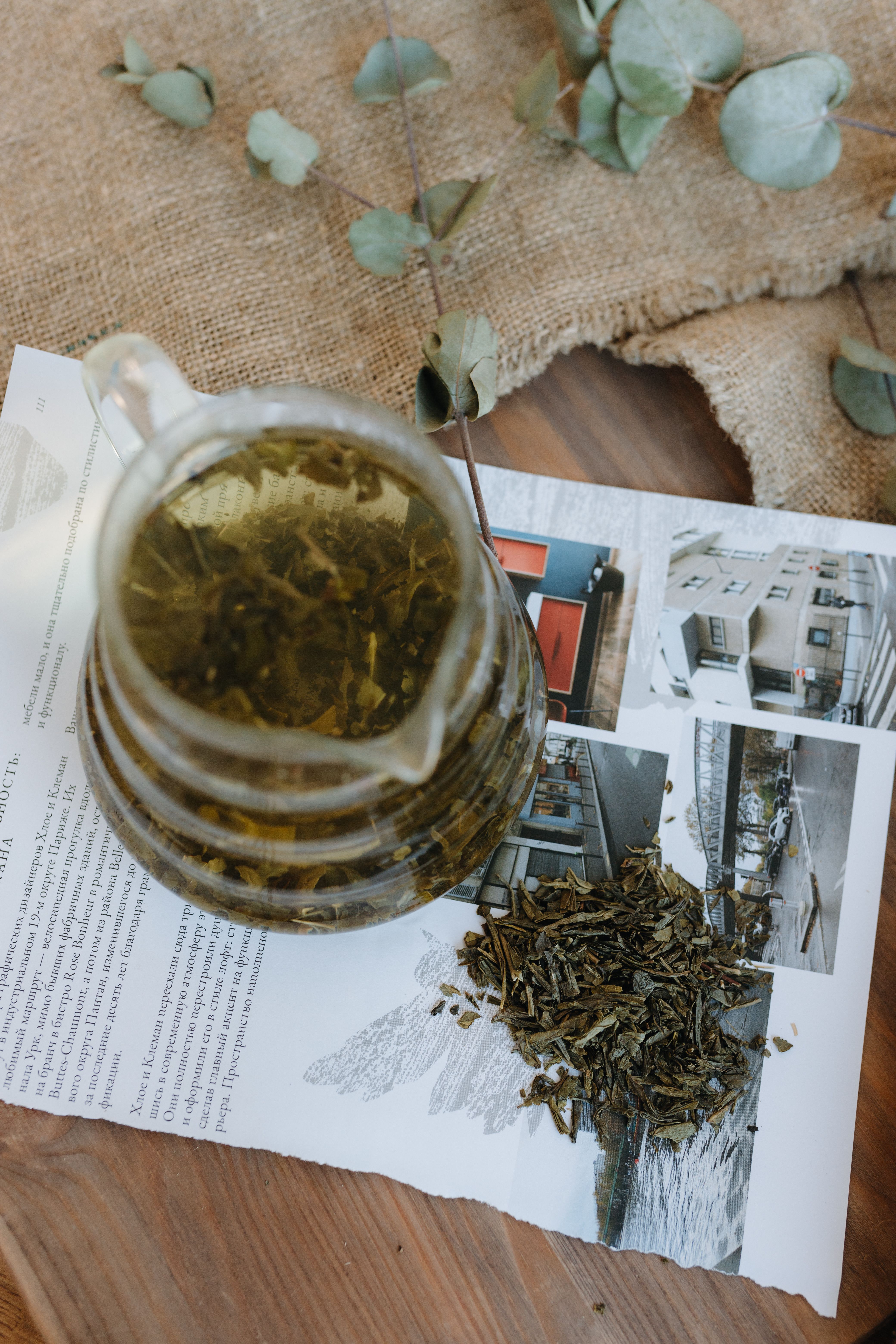 A glass pitcher of green tea atop a rustic table with loose leaves and a magazine.