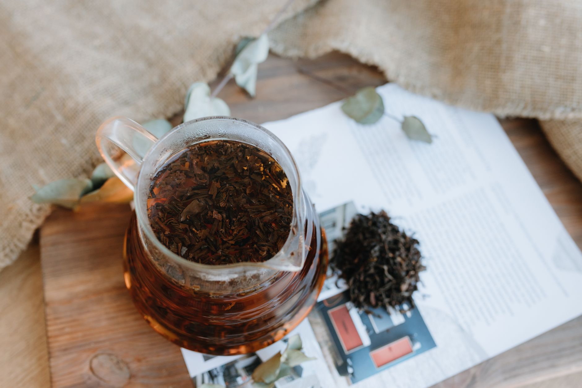 Overhead view of loose leaf tea in a glass pitcher, surrounded by rustic elements on a wooden table.