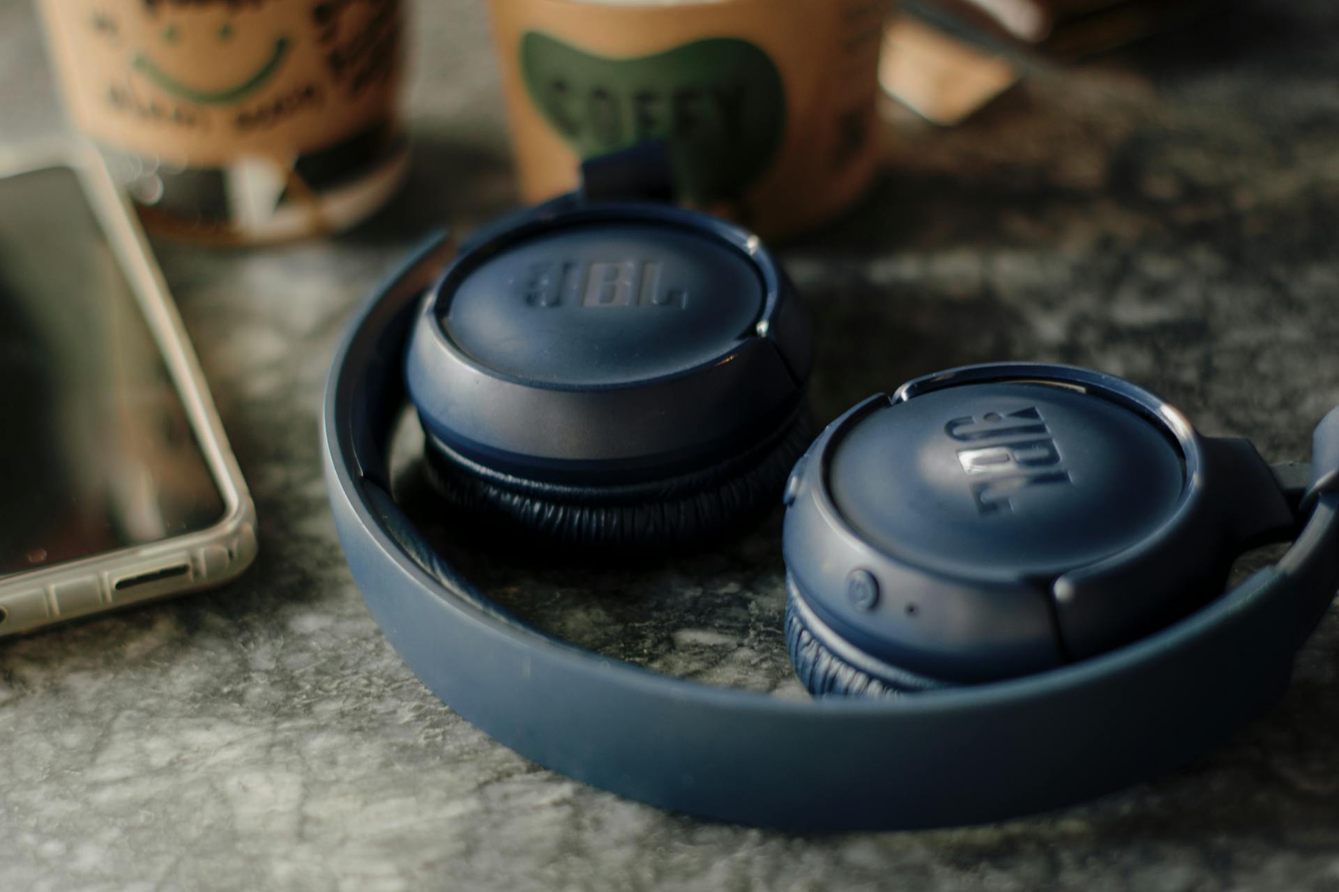 Dark blue JBL wireless headphones beside a smartphone and coffee on a table.