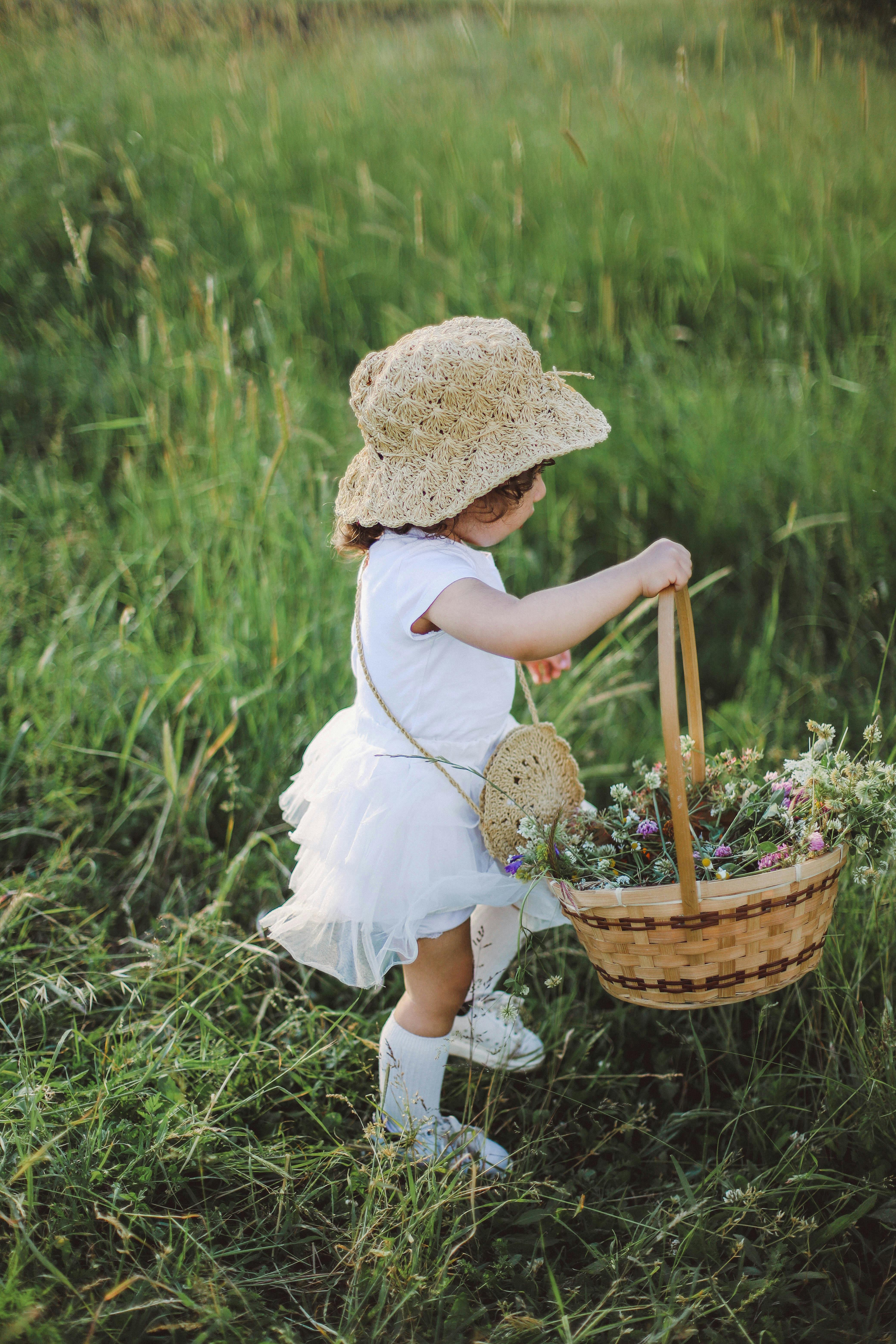 A young girl in a sundress and straw hat carries a basket of flowers in a meadow.