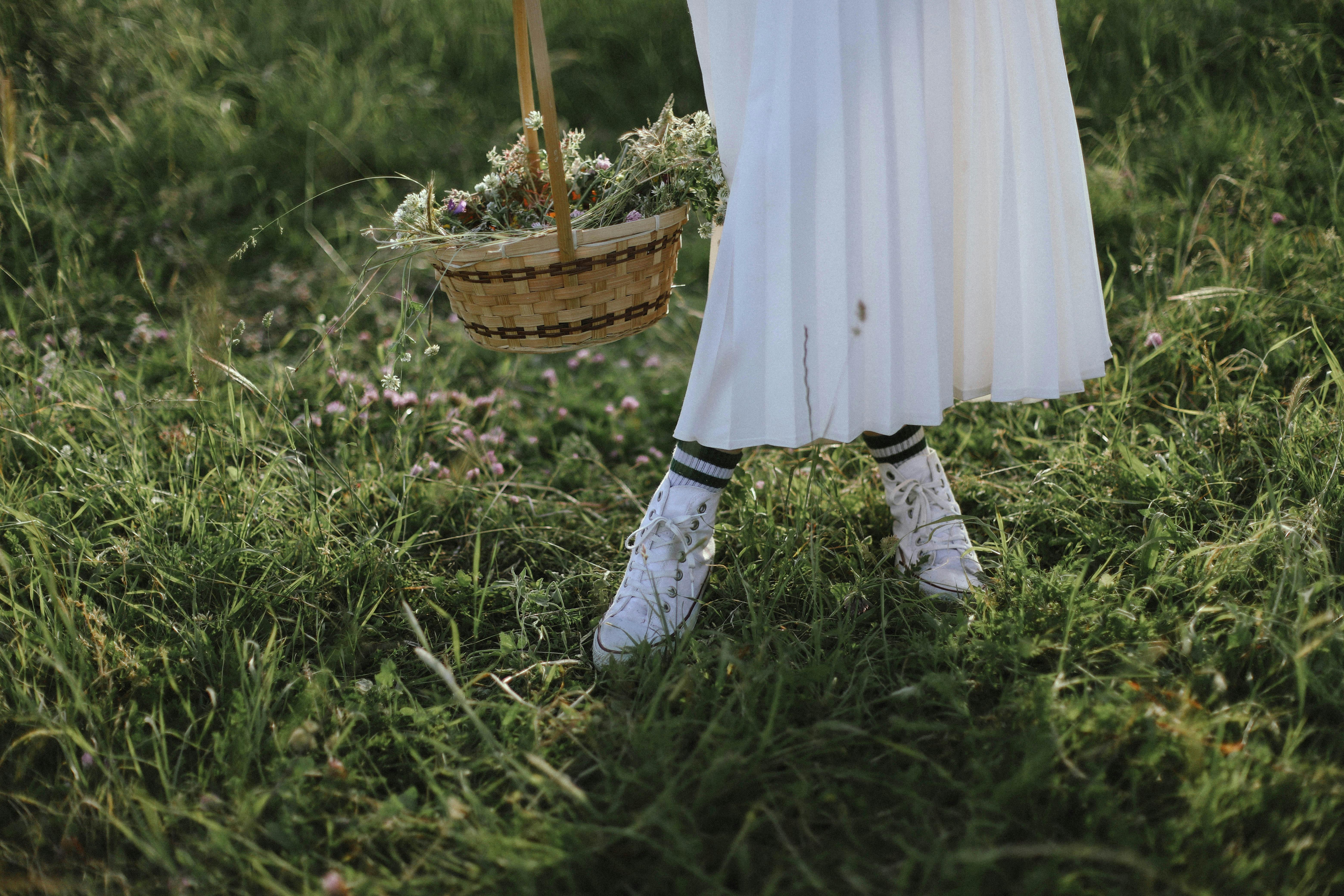 A woman stands in a meadow holding a basket filled with wildflowers, wearing a white dress and shoes.