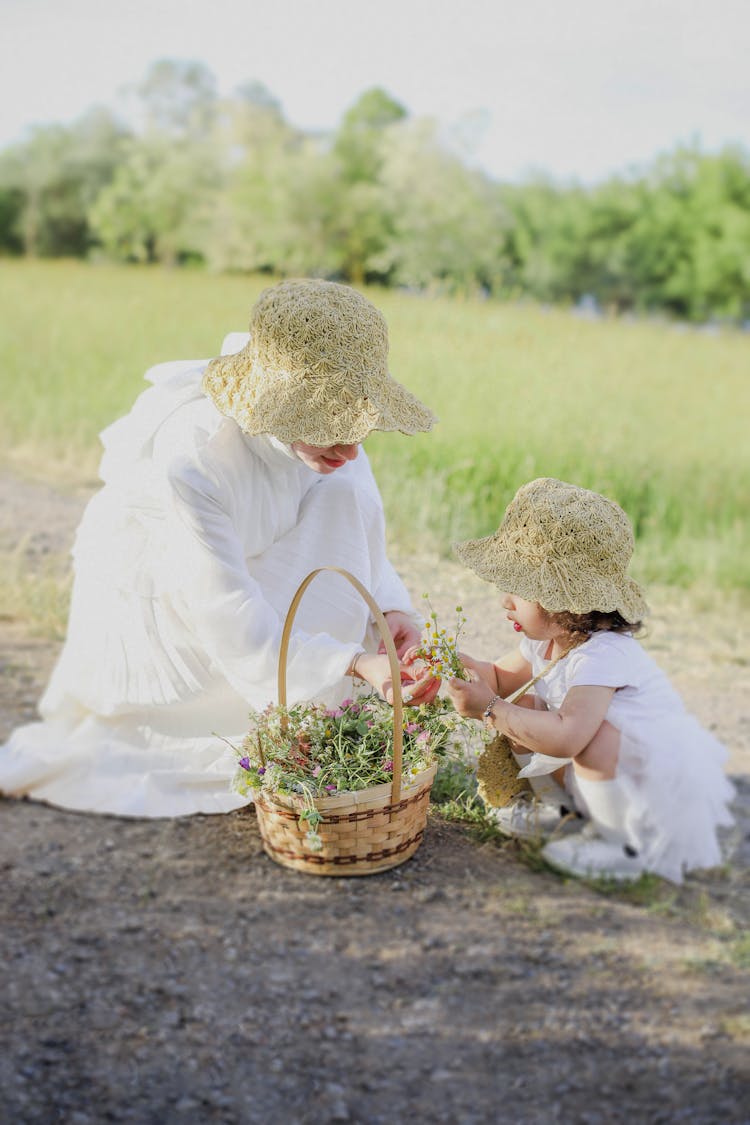 Mother And Daughter In White Dresses And Hats Picking Flowers On A Meadow 