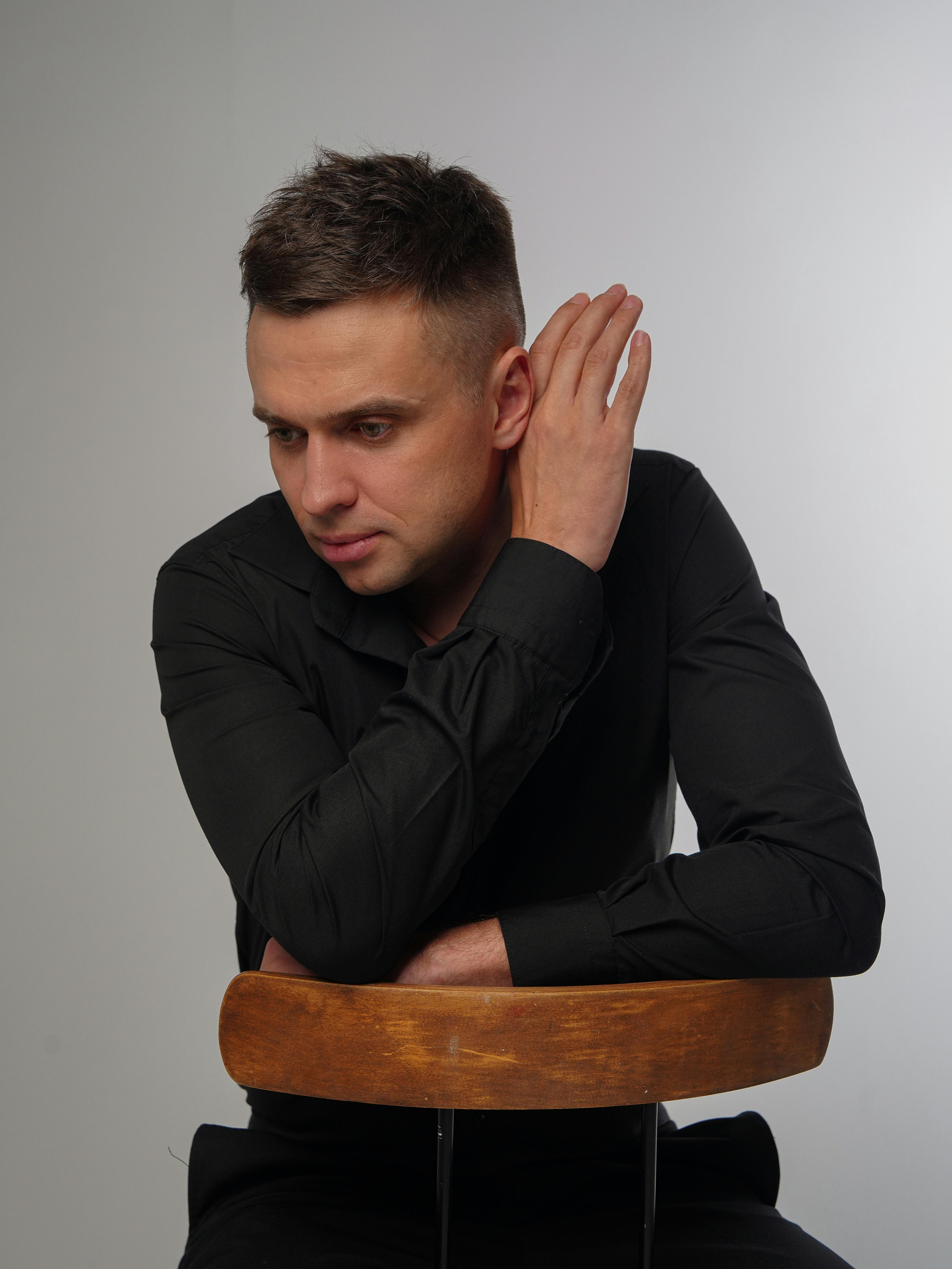 A thoughtful man with short hair in a black shirt sits on a wooden chair, studio setting.