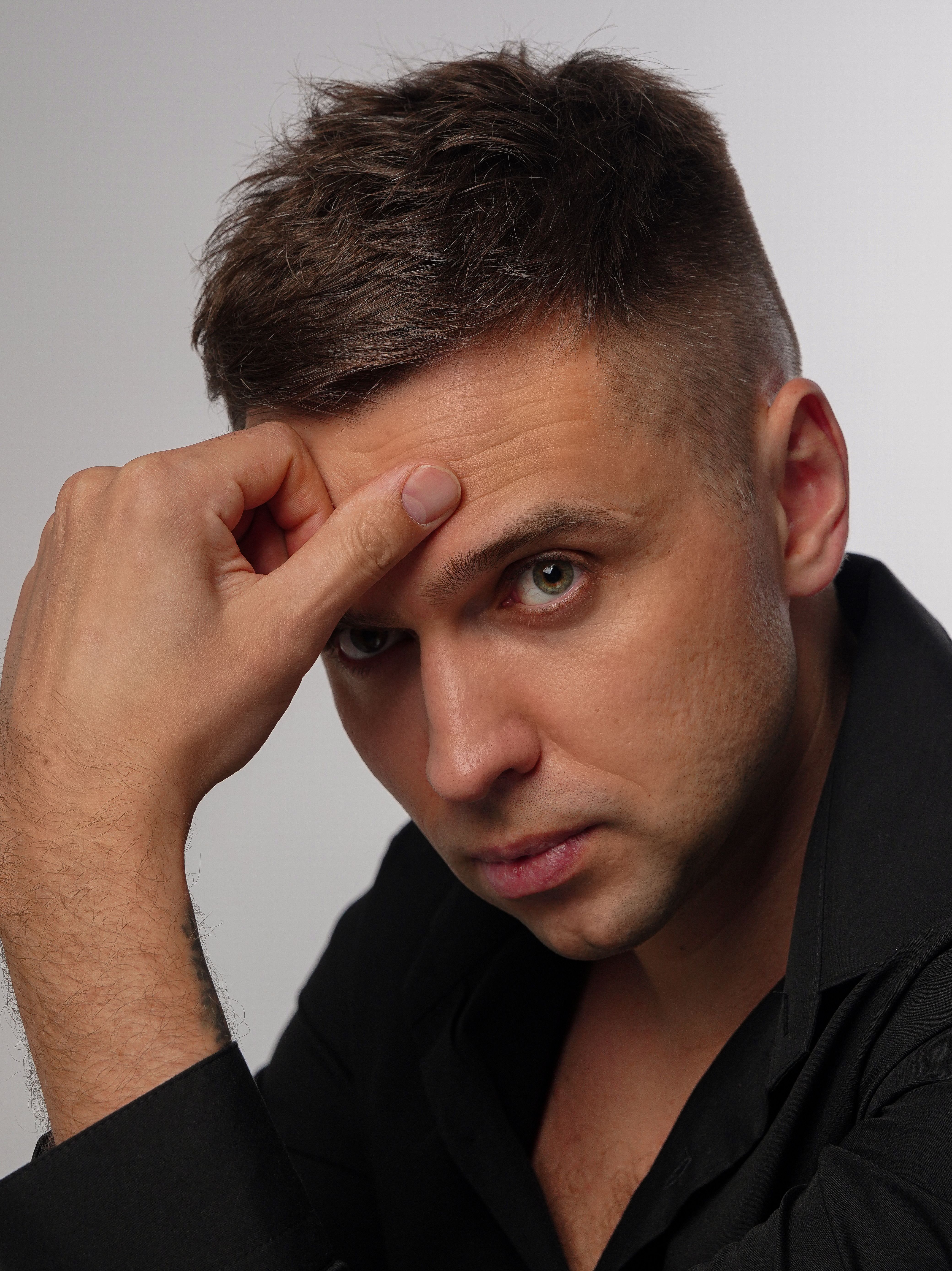 A close-up studio portrait of a young man with a thoughtful pose, hand on forehead, against a white background.