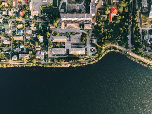 Free Drone capture of a town by the river surrounded by lush greenery and urban architecture. Stock Photo