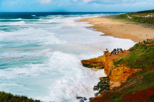 Free Vibrant view of waves crashing on cliffs with beachgoers enjoying a sunny day along the coastline. Stock Photo