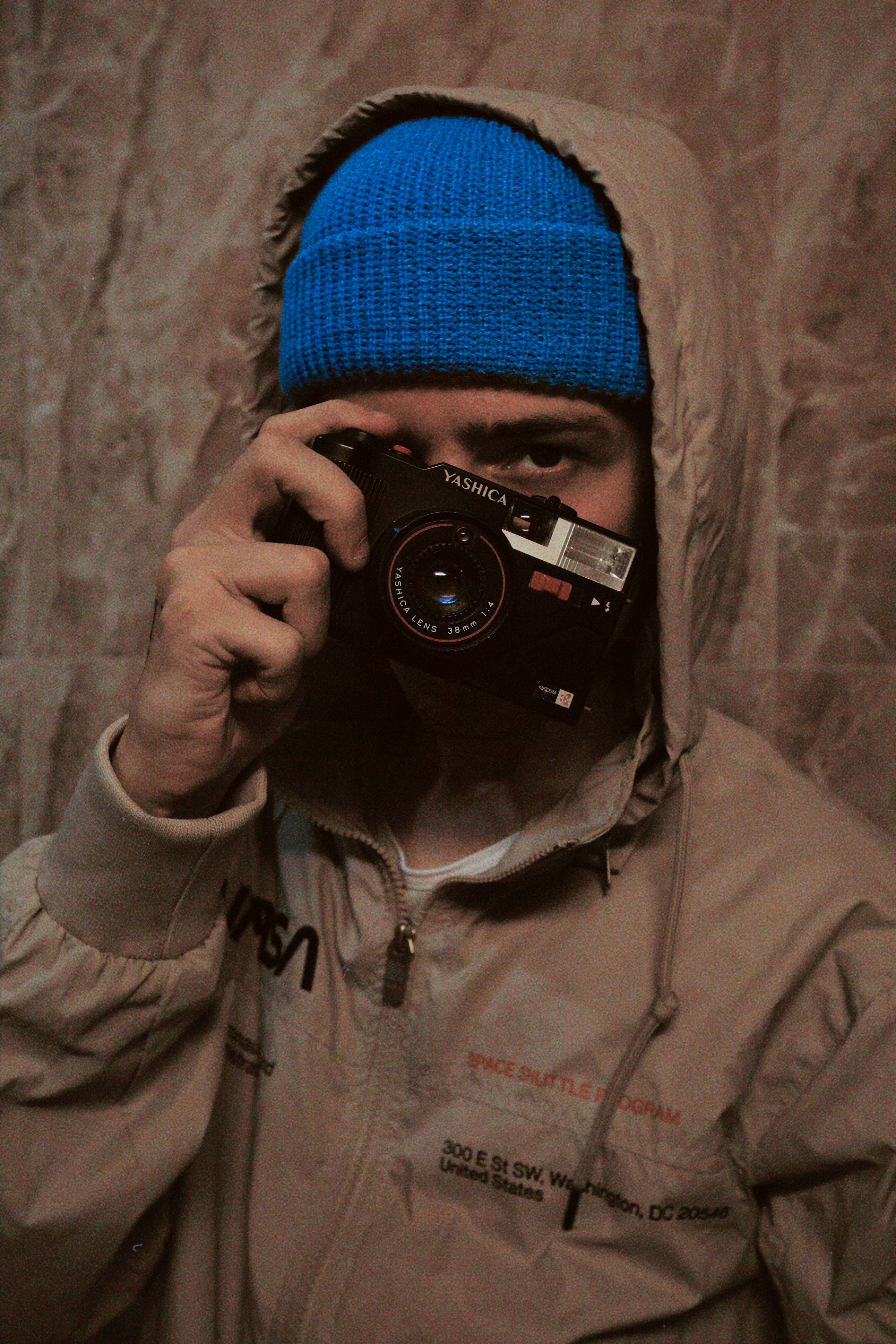 Man in hoodie and blue beanie taking a photo with a vintage camera indoors.