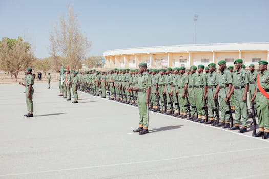 A group of soldiers in green uniforms stands in formation during a sunlit outdoor military parade.