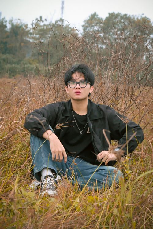Free Portrait of a young man in casual attire sitting in a meadow, wearing glasses and a black jacket. Stock Photo