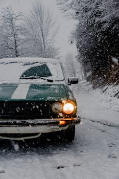 A vintage green car with headlights on, driving through a snowy winter forest road.