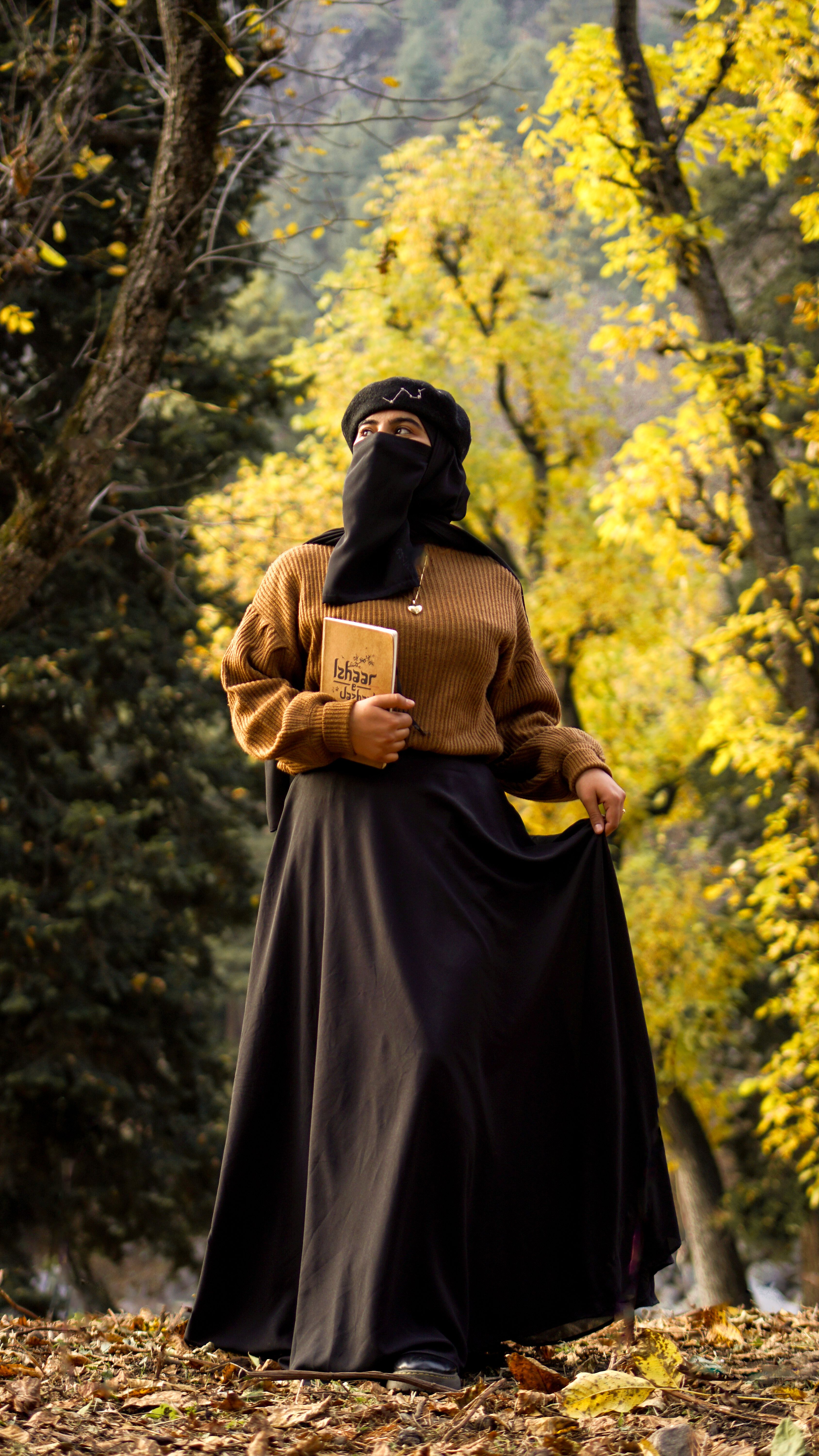Muslim woman in fashionable attire standing in a vibrant autumn forest holding a book.