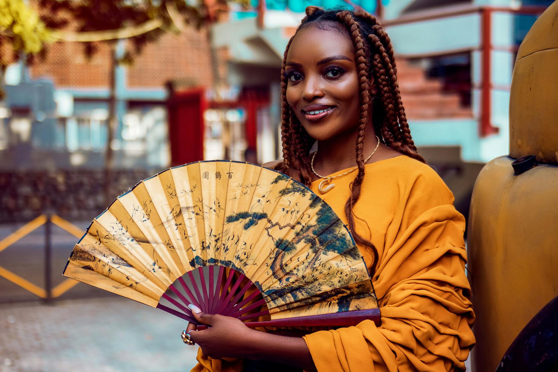 Young African woman in Lagos wearing a yellow dress and holding an Asian fan, embracing cultural diversity.