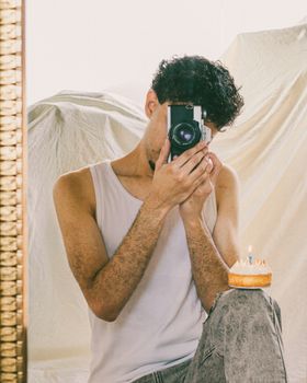 A man in a white tank top sits in front of a mirror holding a camera, capturing a reflection.