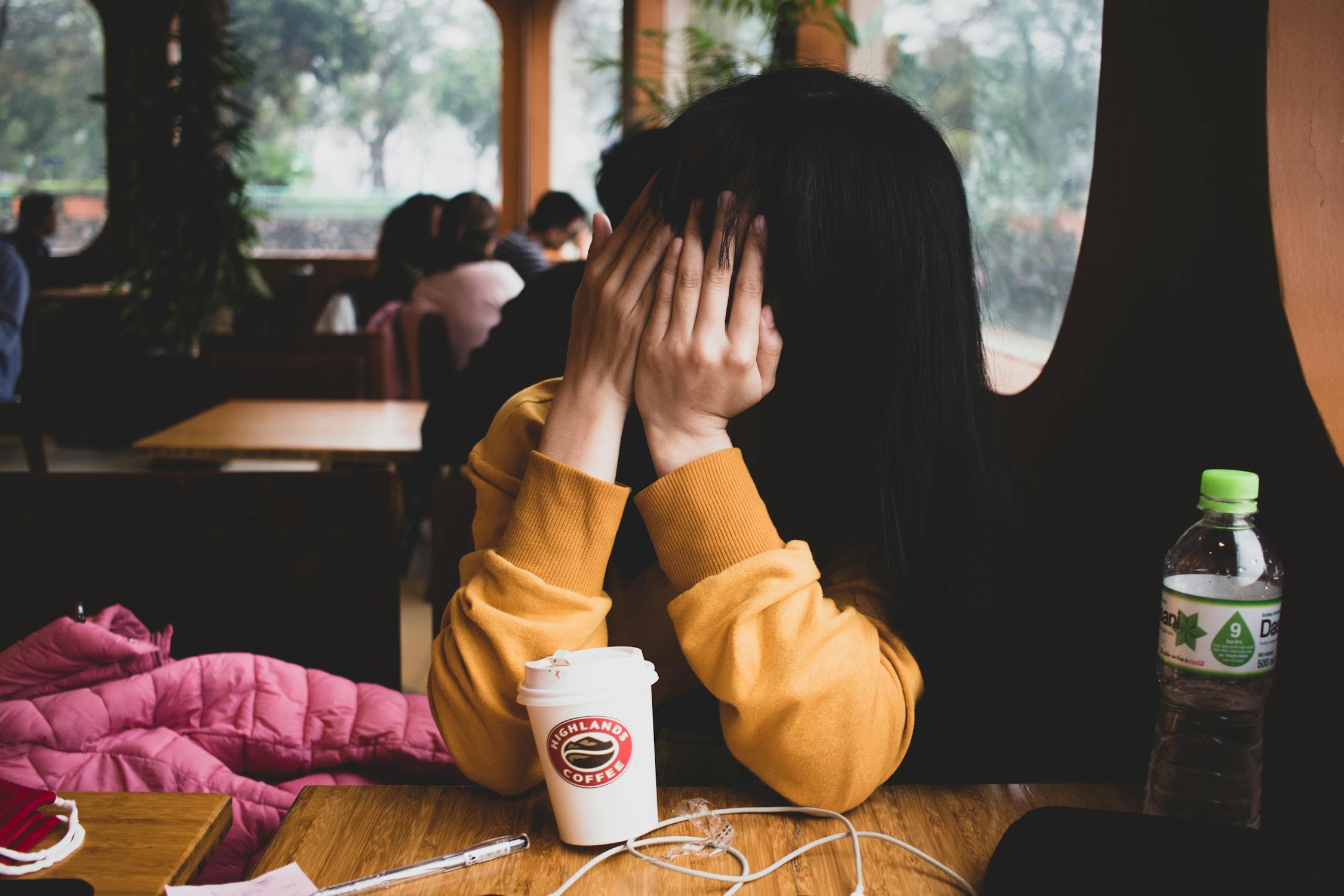 A woman sits at a cafe holding her hands over her face. This represents the painful feelings that come with conflict anxiety. 