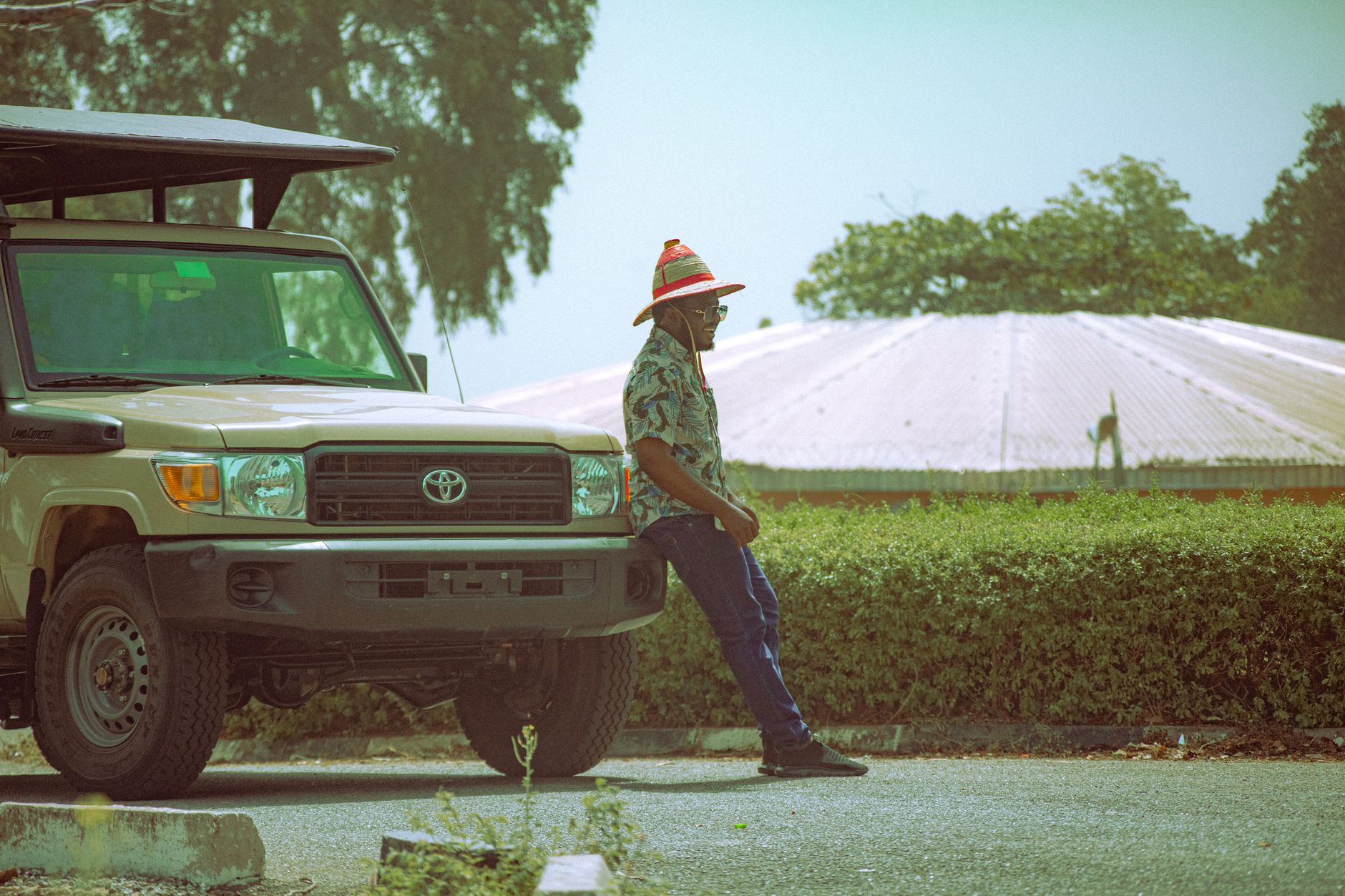 A man in a colorful hat leans against an SUV on a sunny day in Mainamaji, Nigeria.