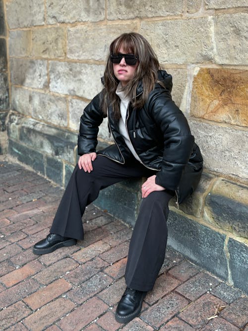 A woman in black leather pants and sunglasses sitting on a brick wall