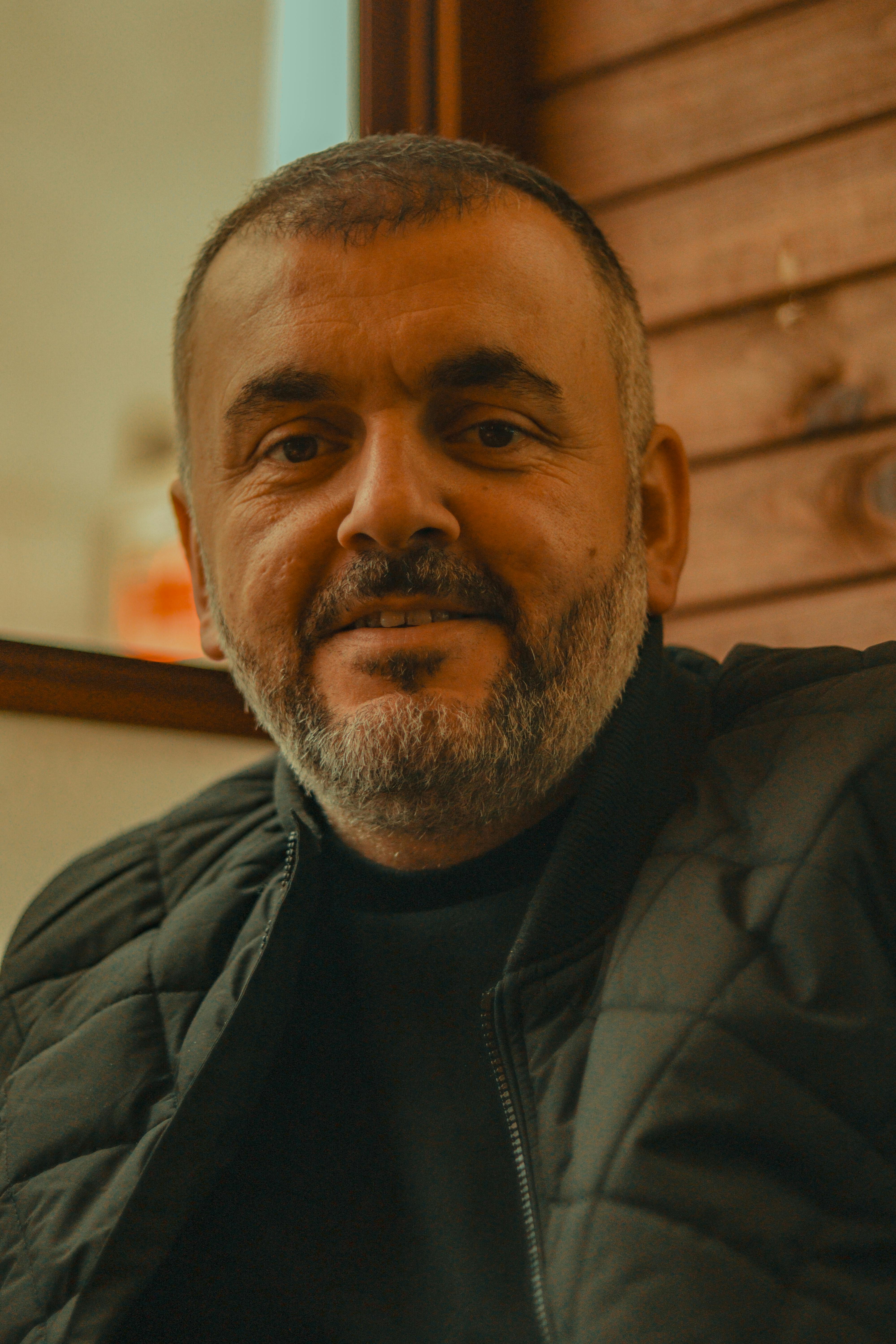 Close-up portrait of a smiling man with a beard wearing a jacket indoors.