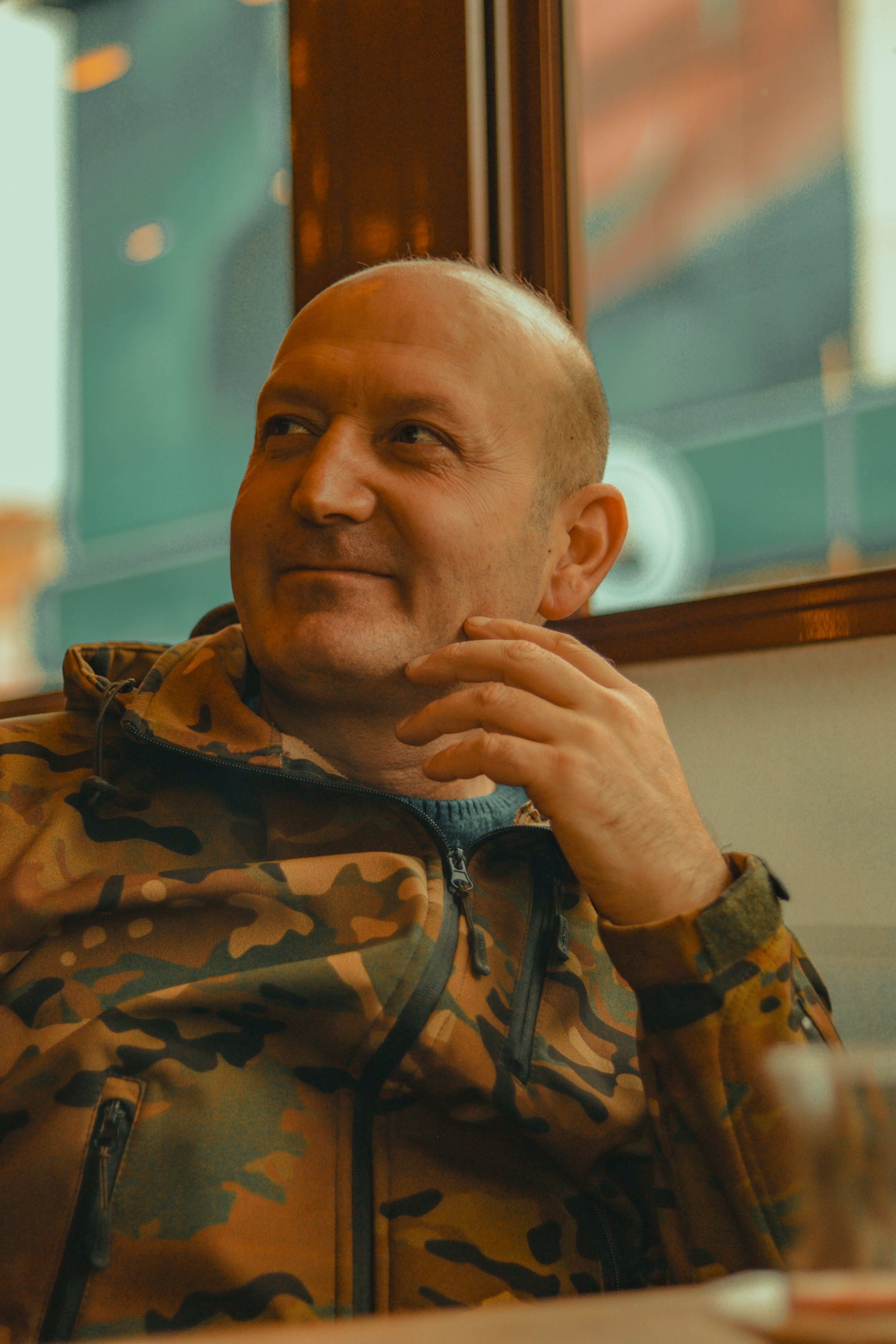 Man casually sitting indoors wearing a camouflage jacket, looking thoughtful.