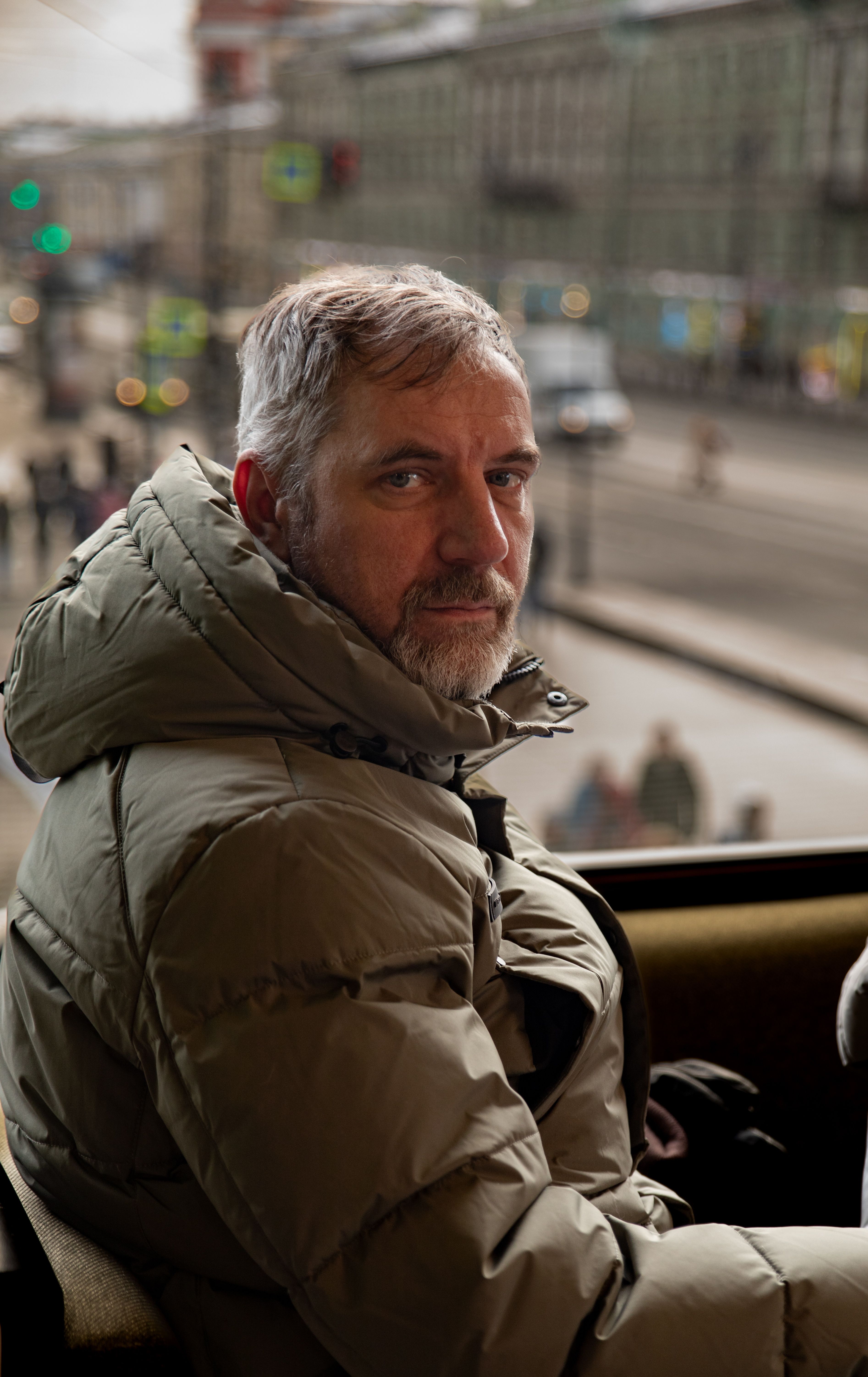 Mature man in a winter jacket sitting by a window with an urban street view.