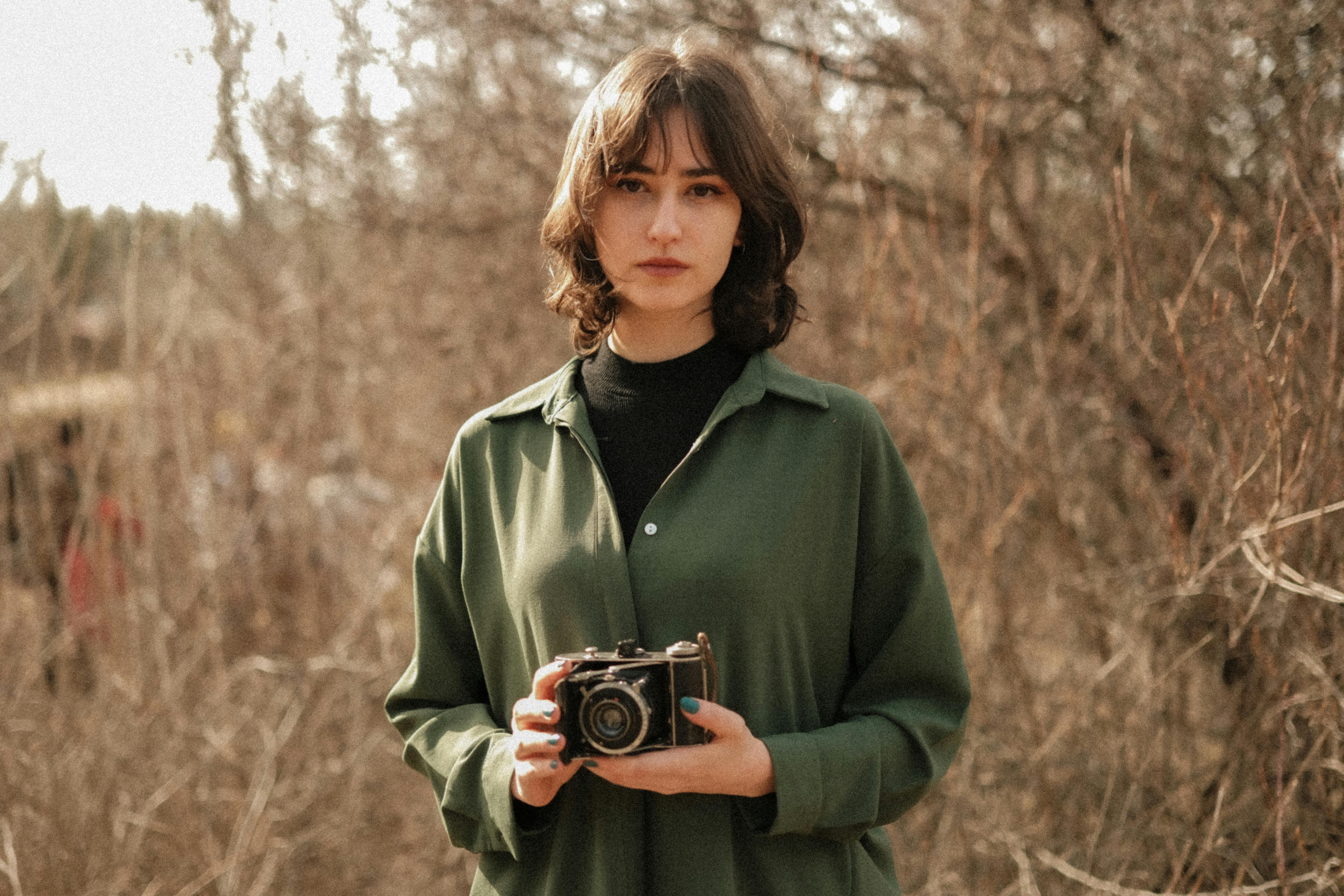 Portrait of a young woman in a green shirt holding a vintage camera in a rural setting.