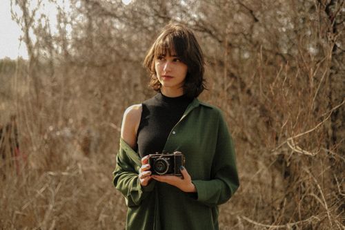 Free A fashionable woman holding a vintage camera, outdoors in a natural setting with dry foliage. Stock Photo