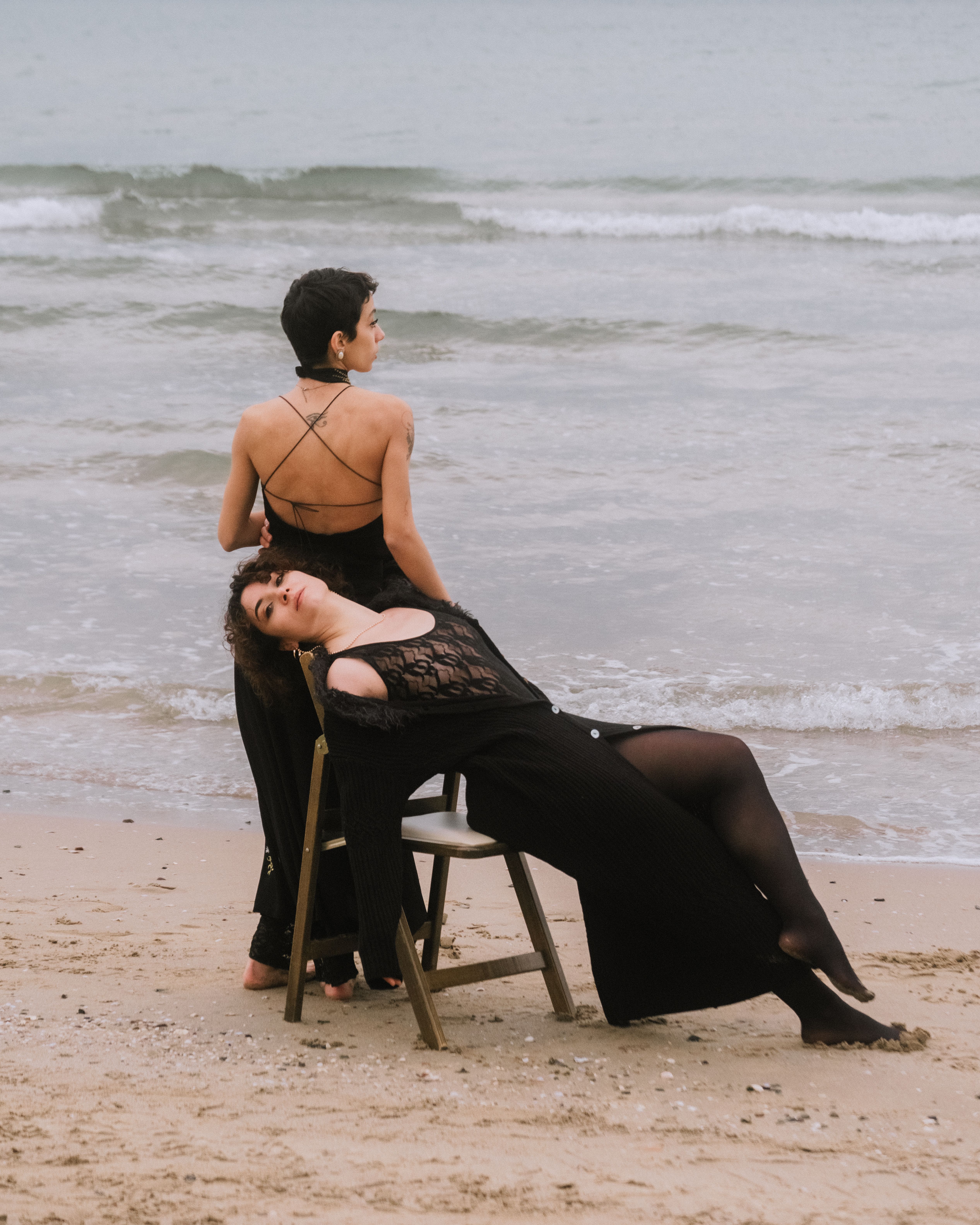 Two women in black dresses enjoying a peaceful moment on a sandy beach with gentle waves.