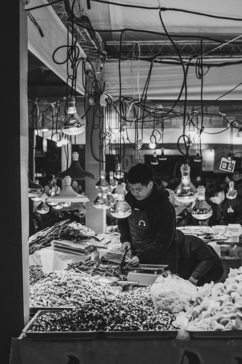 Free Black and white photo of a vendor working at a bustling market stall under hanging lights. Stock Photo